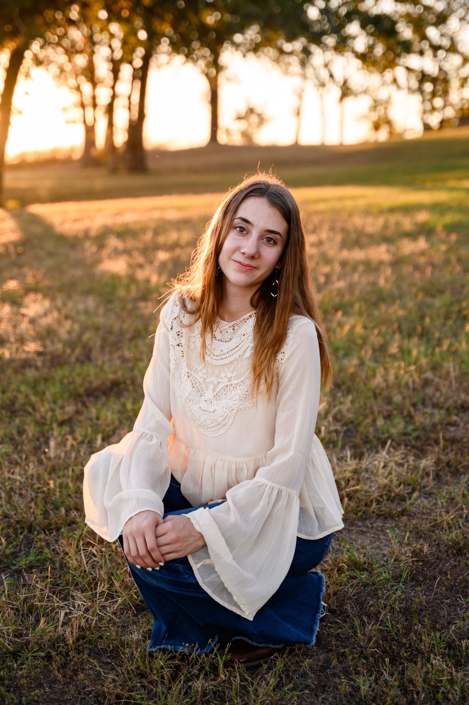 A senior girl in a white lace blouse with bell sleeves sits in a grassy field during golden hour sunset at Big Hill Lake.