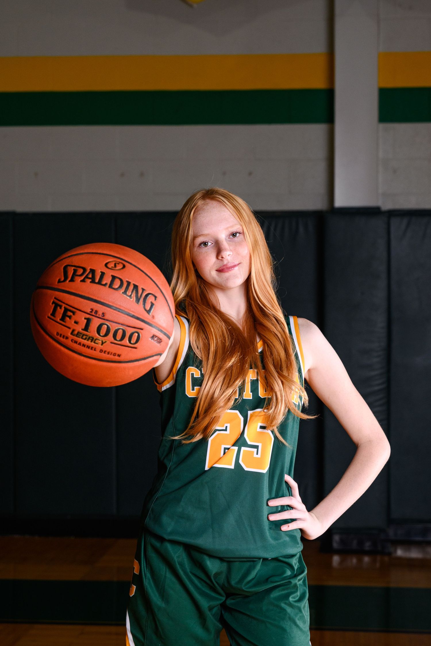 Senior basketball player in green and gold uniform number 25 poses with Spalding TF-1000 ball in gymnasium.