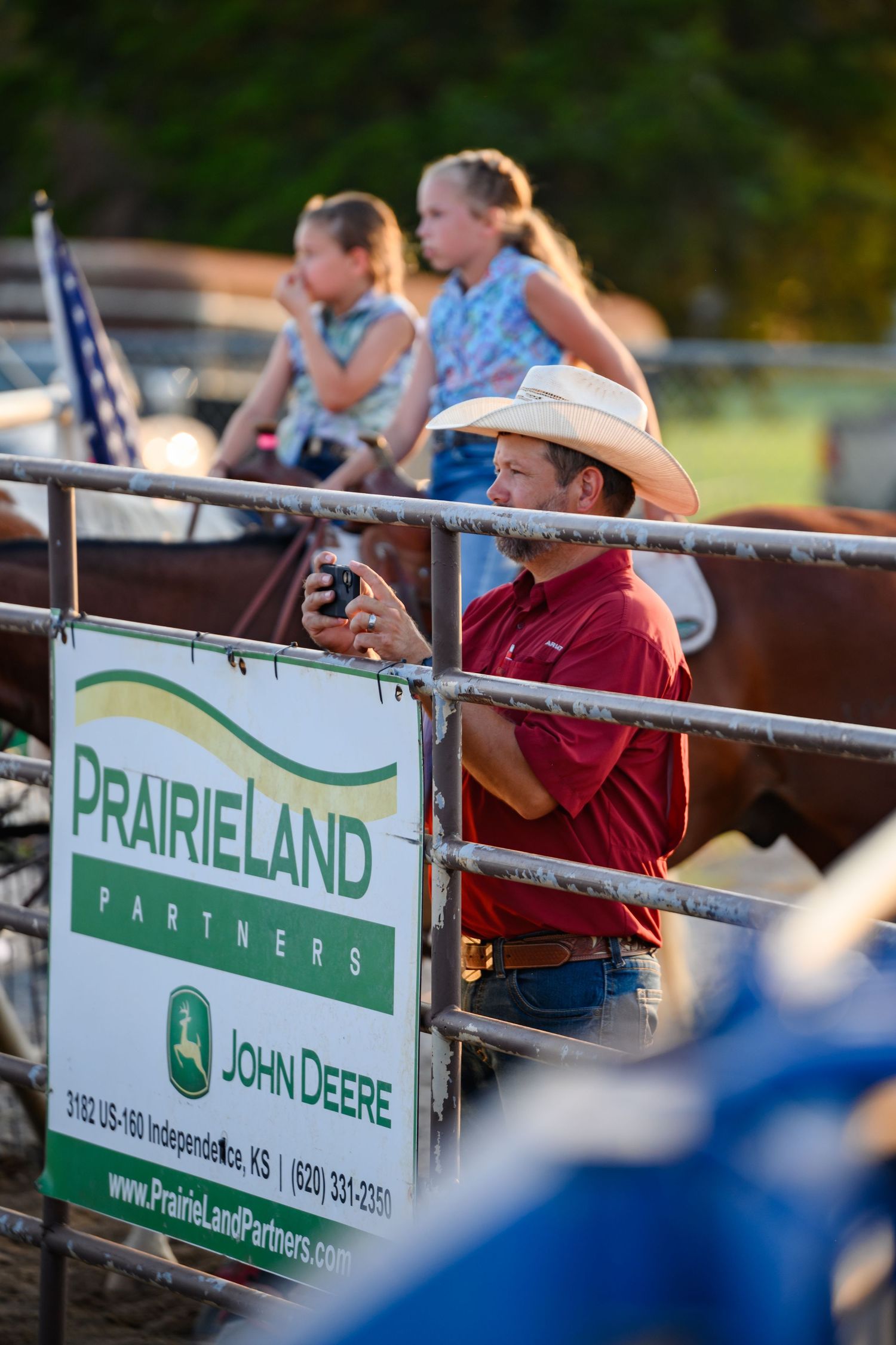John Deere equipment dealer banner displayed at outdoor event with spectators viewing from behind fence railing.