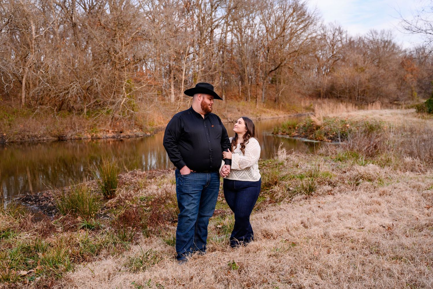 A couple stands together on a grassy shoreline near a pond during an autumn engagement photoshoot.