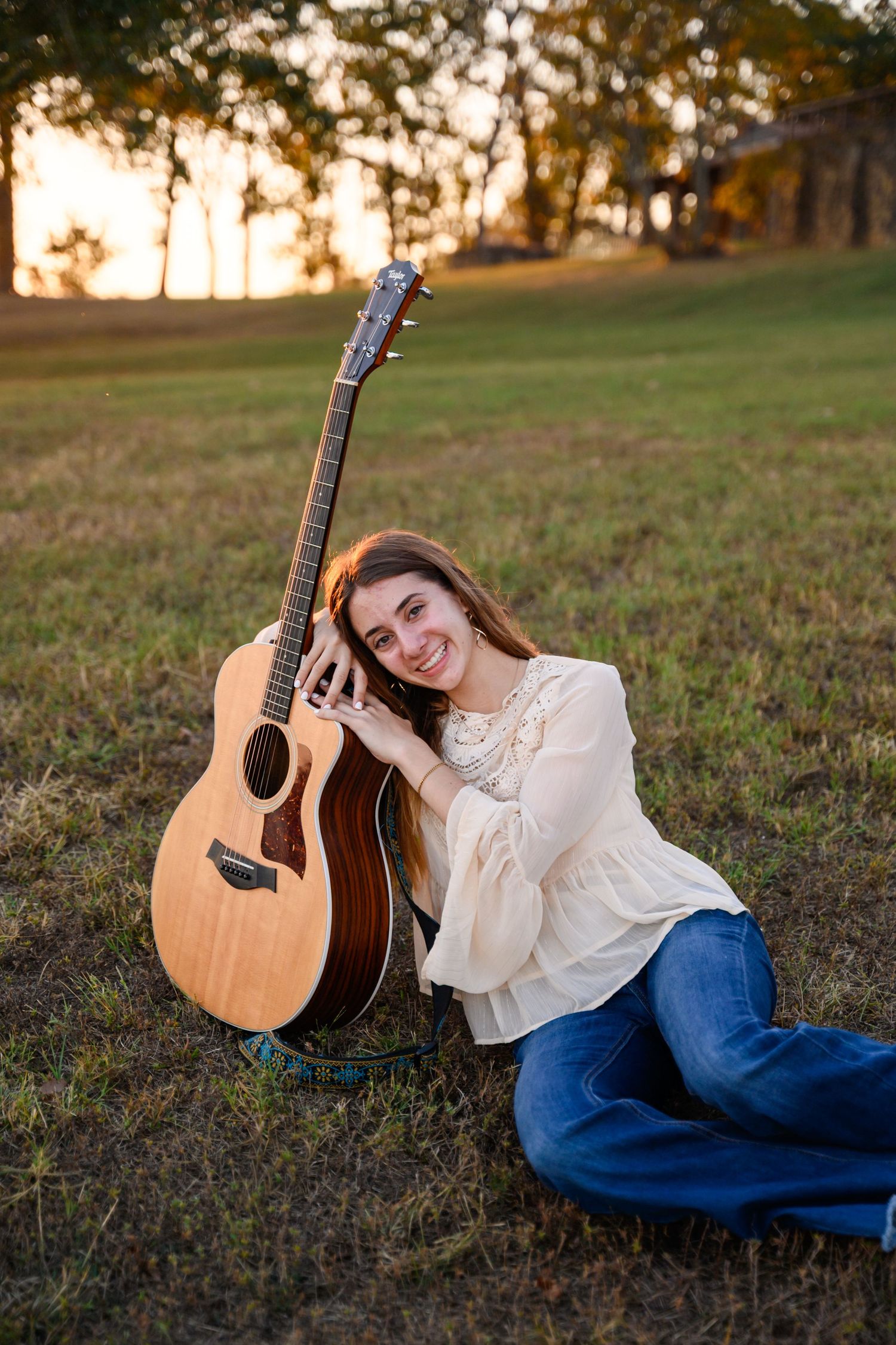 A senior girl in a white blouse and jeans poses with an acoustic guitar on grassy ground at sunset.
