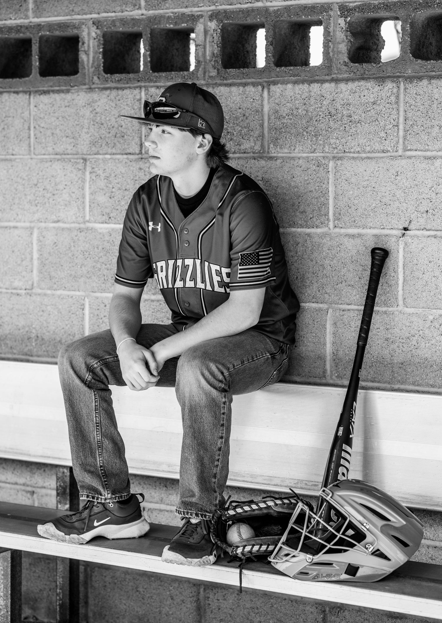 A senior baseball player sits alone in the dugout with a bat and glove on a bench.