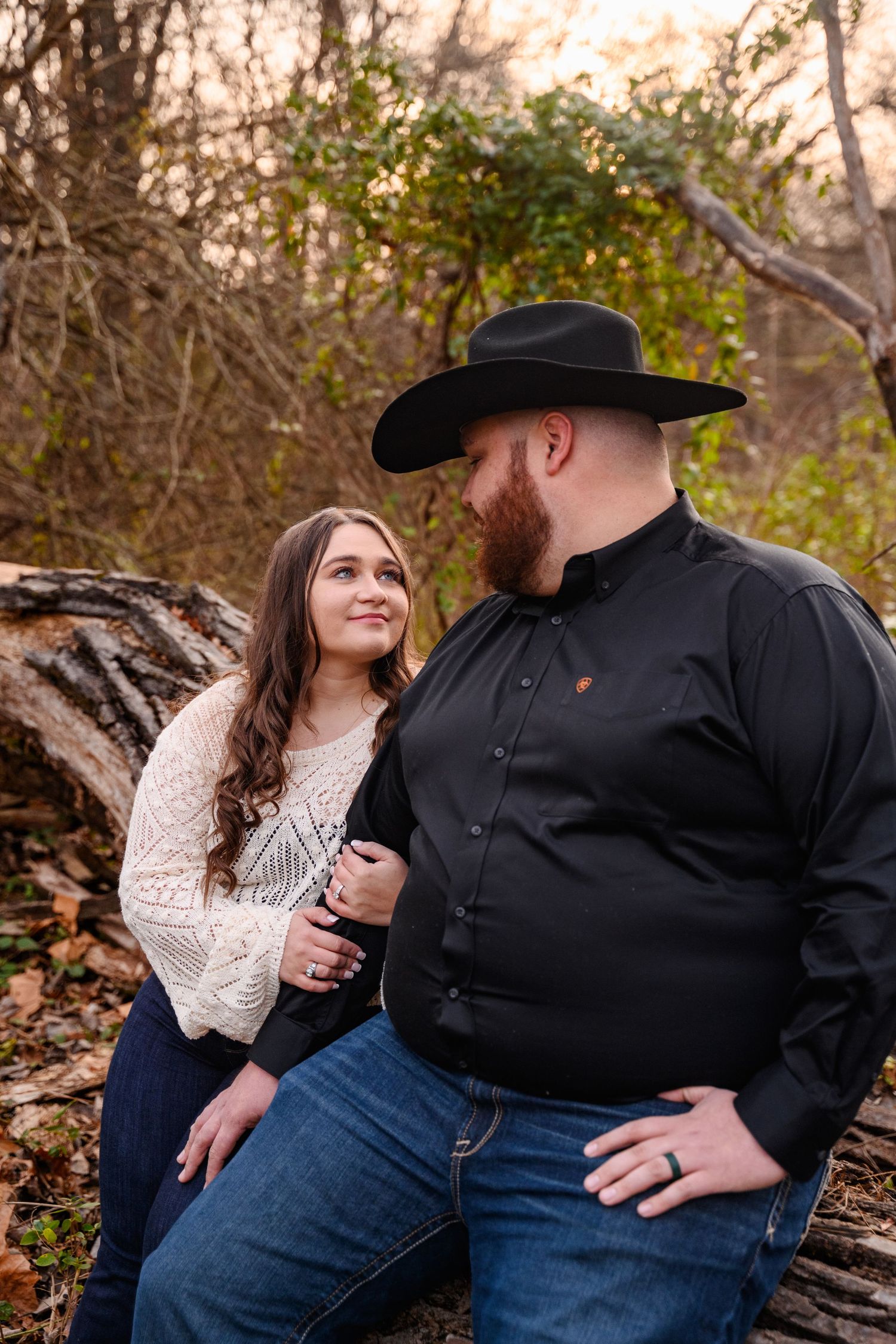 A couple sits together on a fallen log during an autumn photoshoot, with him wearing a black cowboy hat and her in a cream sweater.