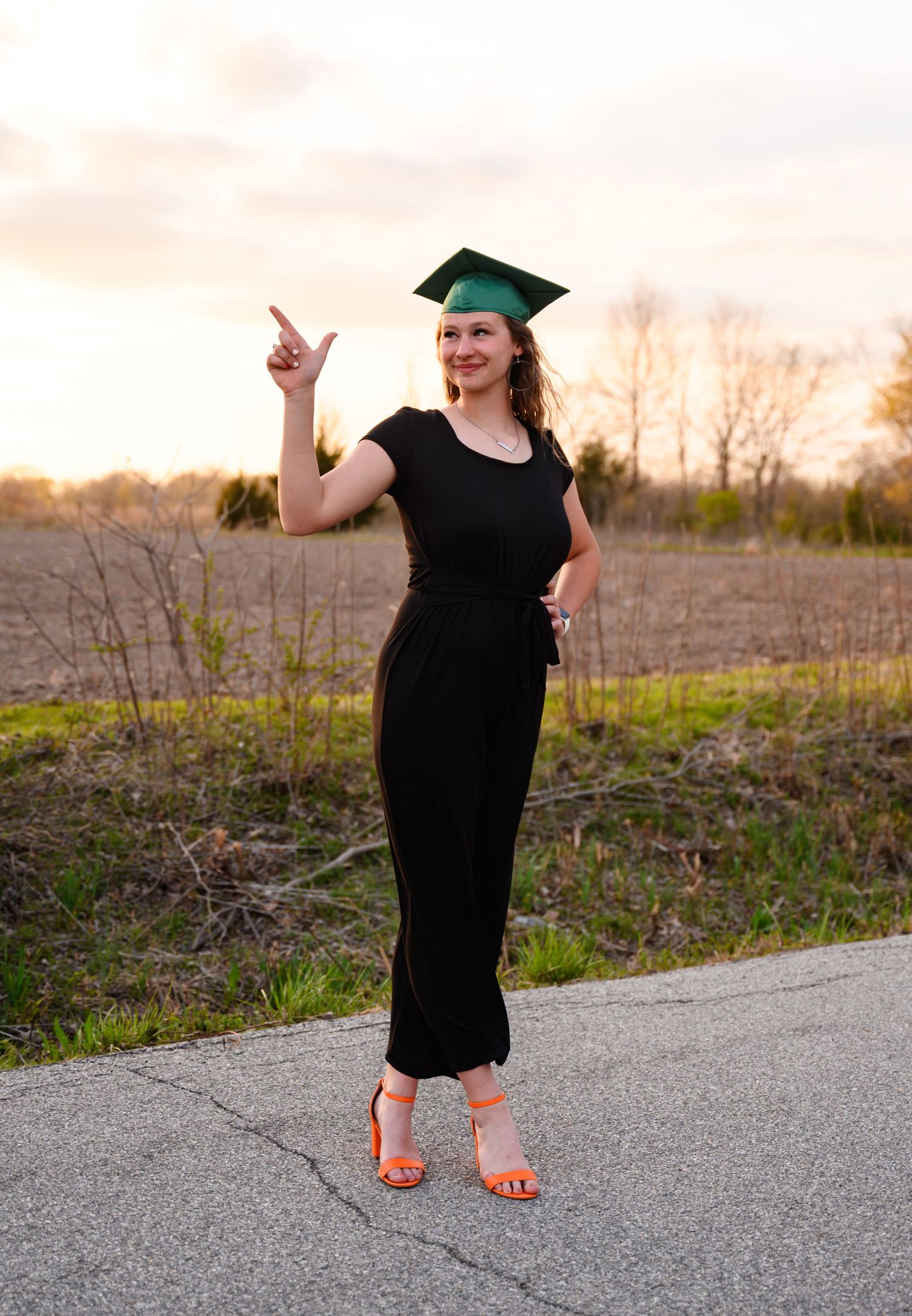 A graduate in a black dress and cap stands on a rural road in Southeast Kansas at sunset, showing her OSU pride.