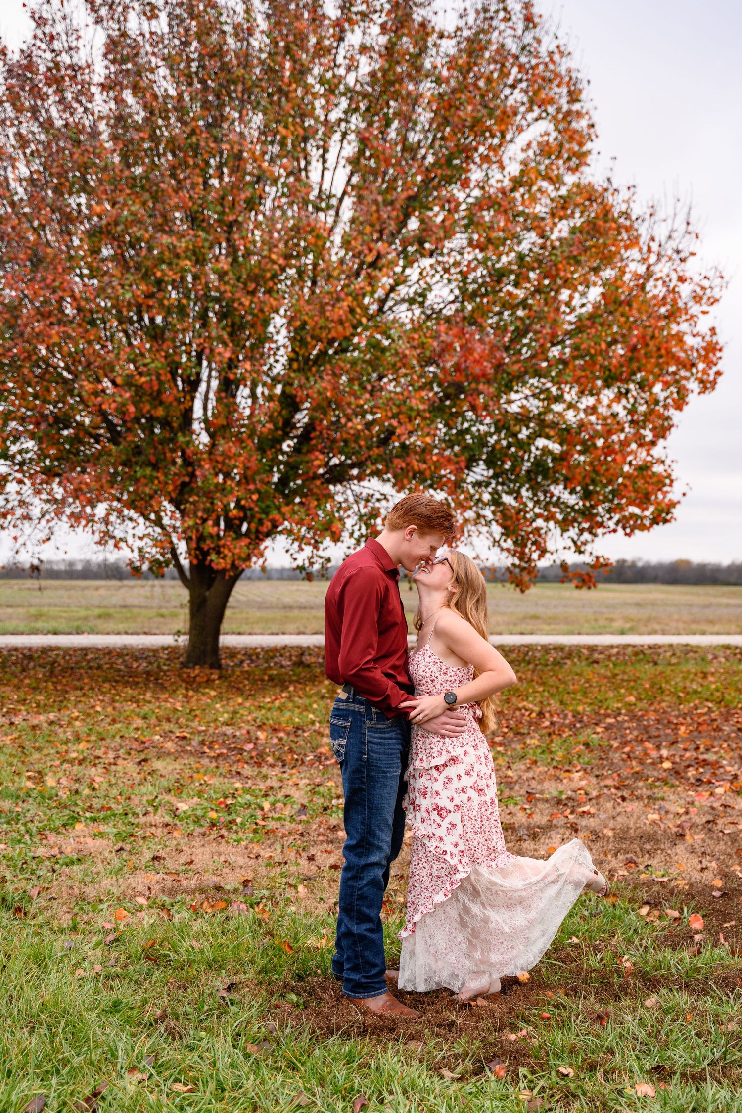 A couple shares romantic moments under a vibrant orange autumn tree in a rural field in Southeast Kansas.