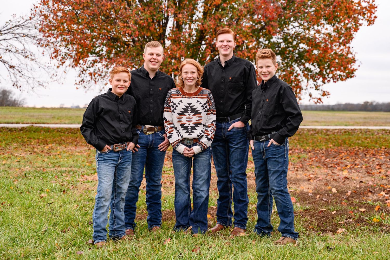 A family stands together outdoors in autumn with vibrant orange fall foliage and trees in the background.