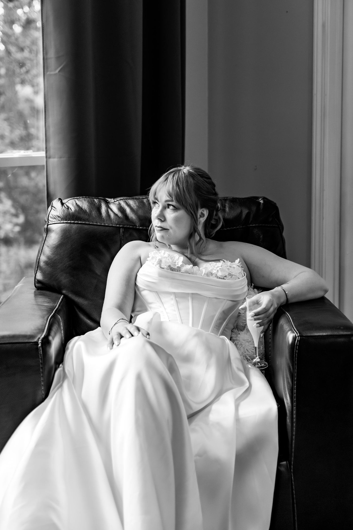 A bride in a white dress reclining on a couch near a window, sipping champagne.