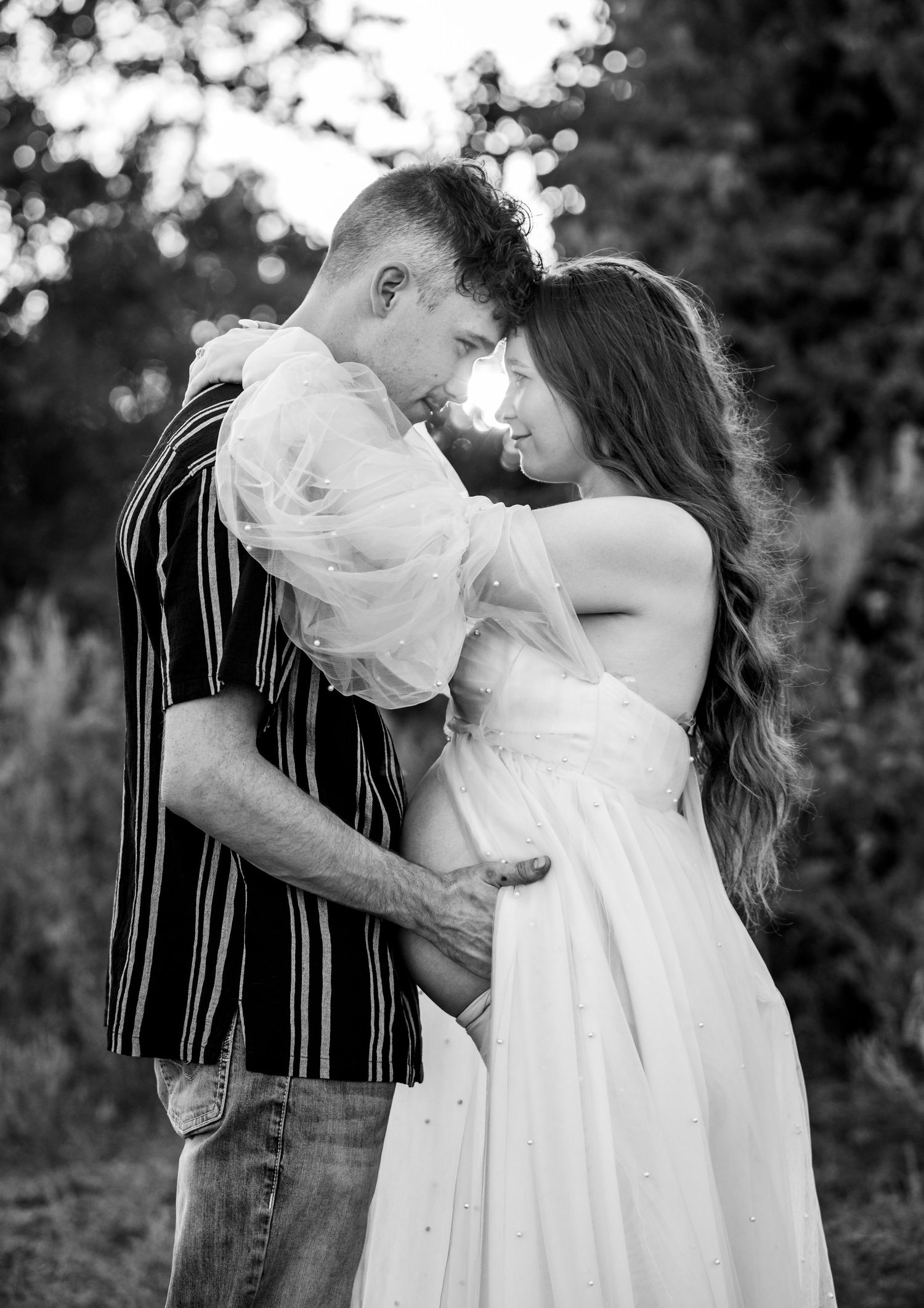 An expecting couple holding their foreheads together, looking into each other's eyes as the sunset peeks through.