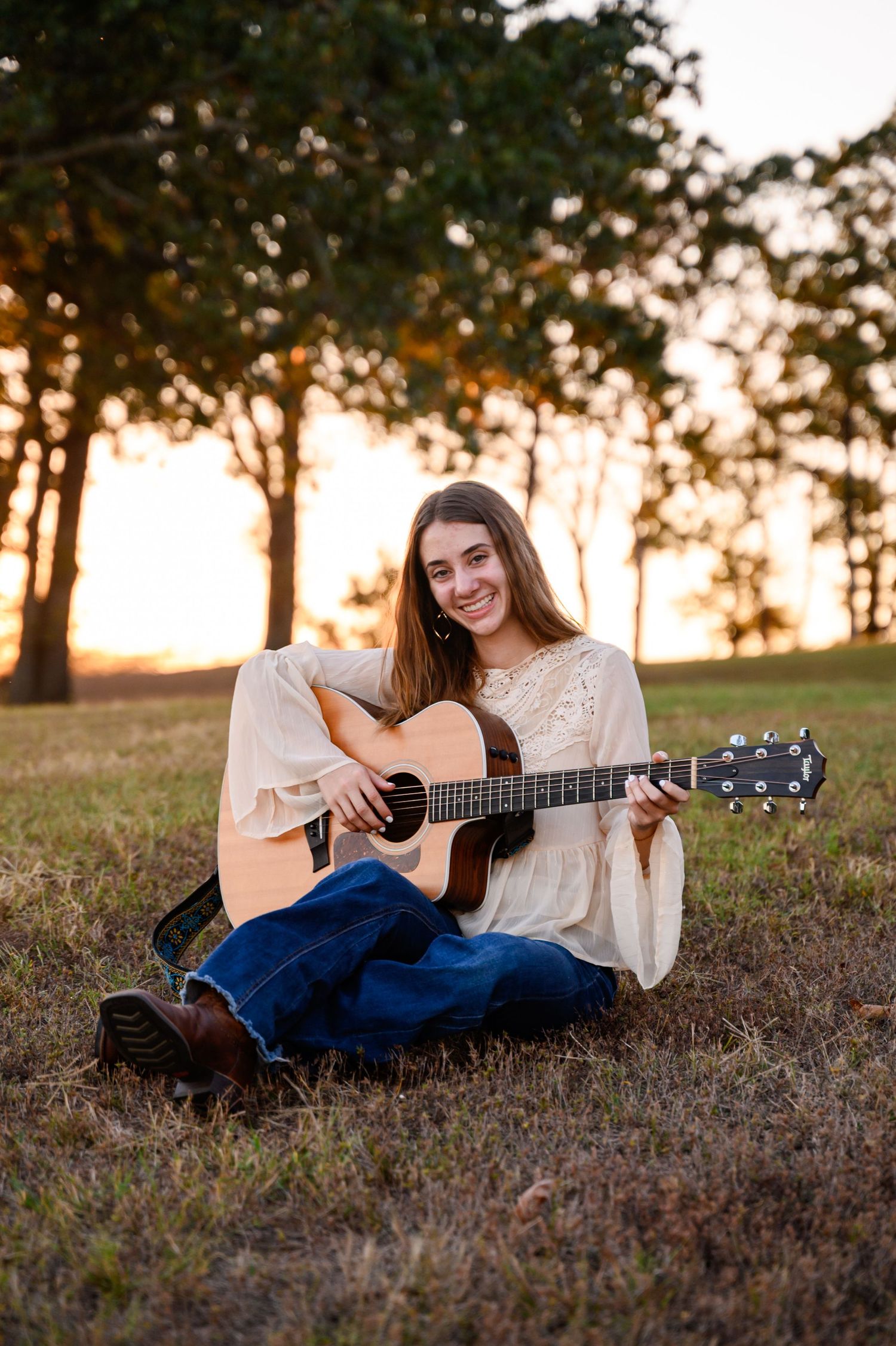 A young musician, a senior girl, in a white blouse and jeans, plays acoustic guitar while sitting on a grassy hill at sunset.