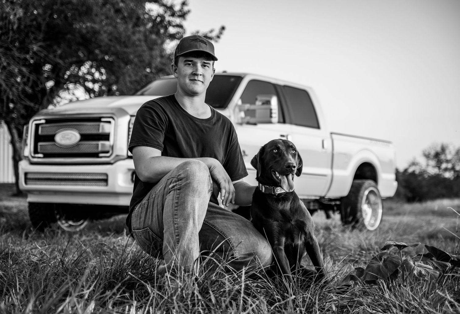 A senior boy and his dog are sitting together in front of his truck in an open field.