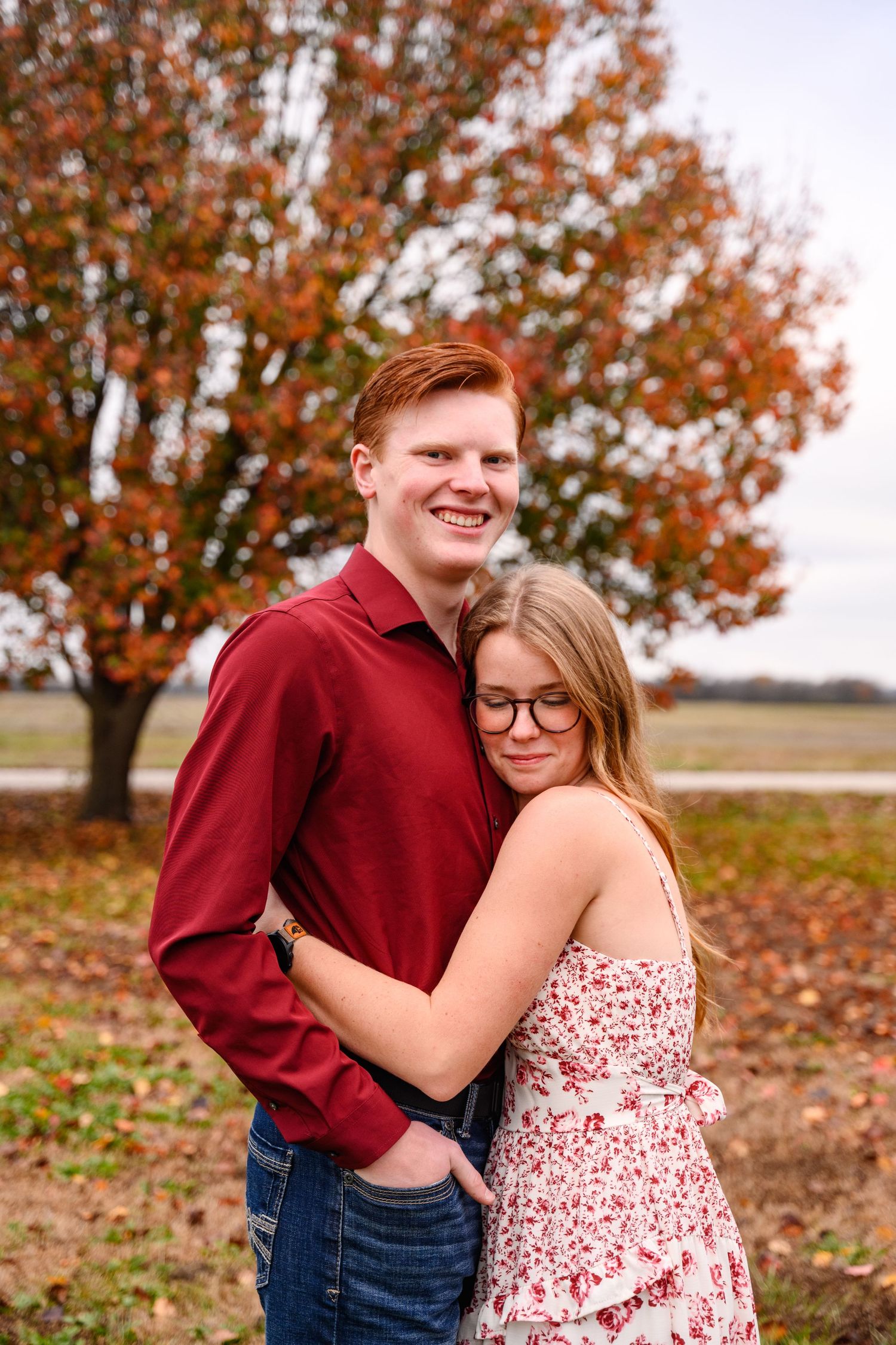 A couple embraces outdoors during autumn with red and orange fall foliage in the background.