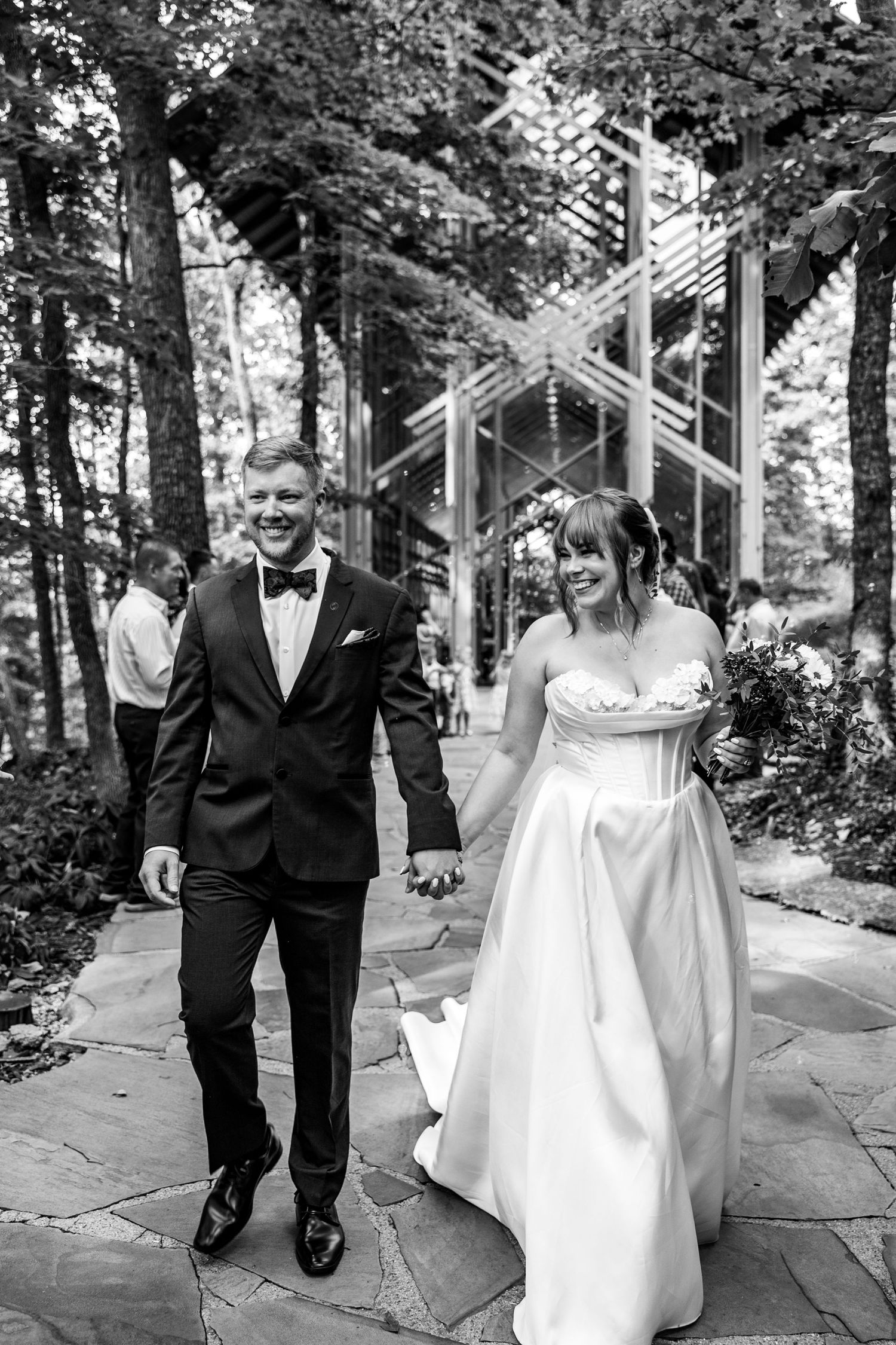 A couple walks together along a stone path in a wooded area, with the bride wearing a flowing white gown.