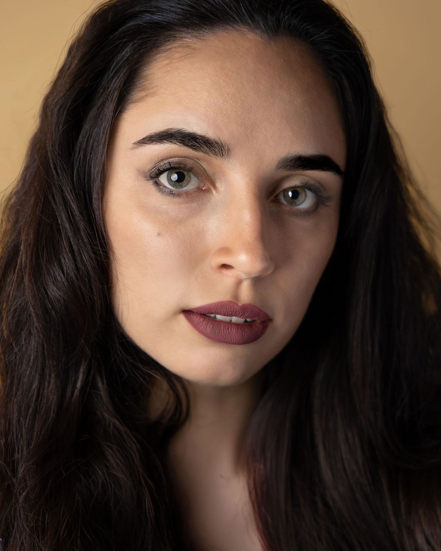 Close up portrait with dark wavy hair, burgundy lipstick and striking eyes against warm brown background.