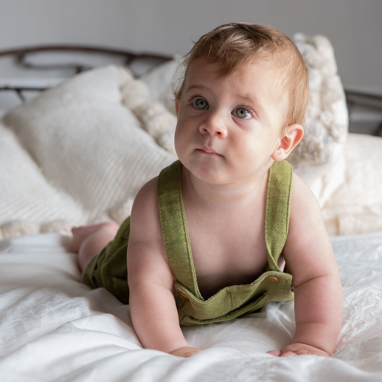 Baby wearing green overalls lies on white bedding in sequence of gentle expressive poses.