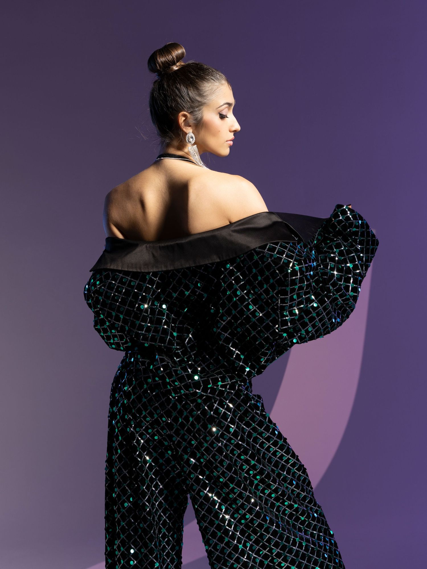 A fashion model poses in a dramatic black sequined dress with sculptural ruffles against a purple backdrop.