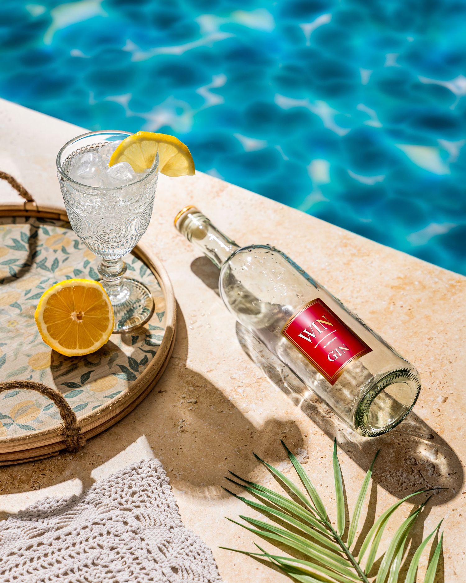 Empty glass bottle and cocktail with orange slice sitting poolside on a sunny day with turquoise blue water.
