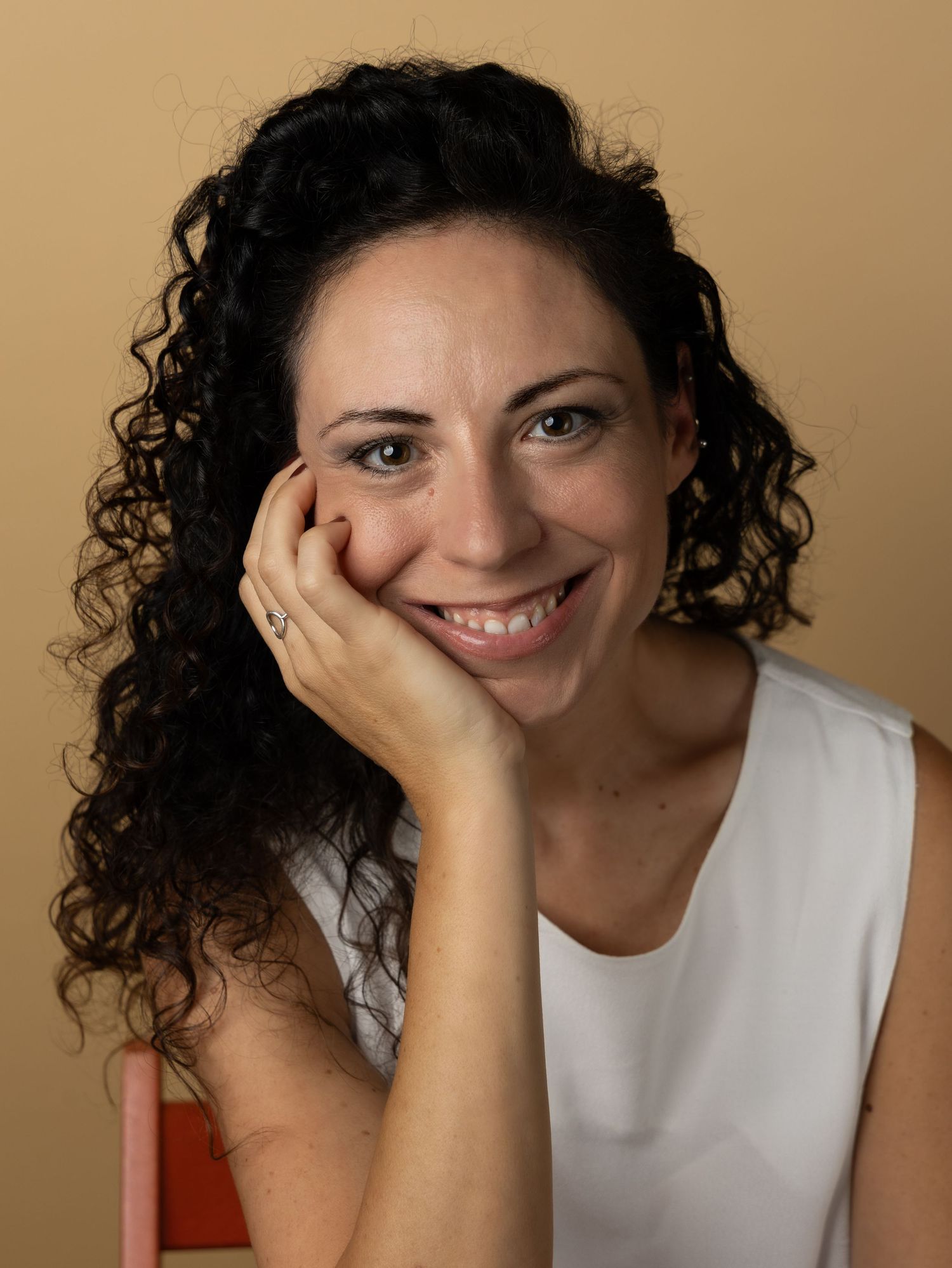 Portrait shot of a person with curly dark hair in a white sleeveless top smiling warmly while resting their chin on their hand.