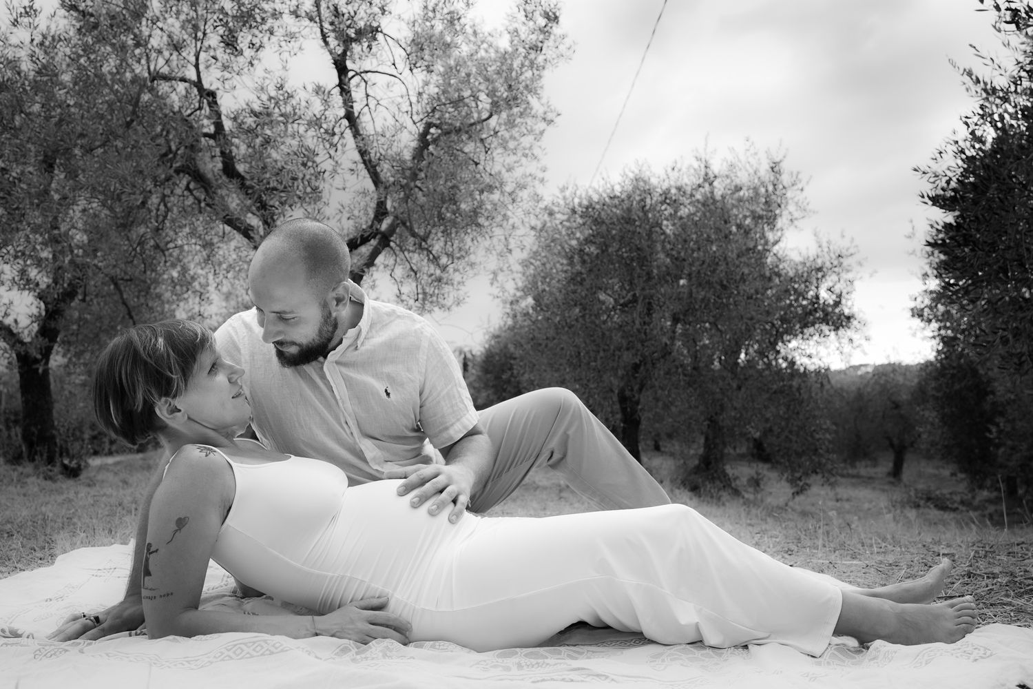 Black and white romantic portrait of a couple laying together in an olive grove on the ground.