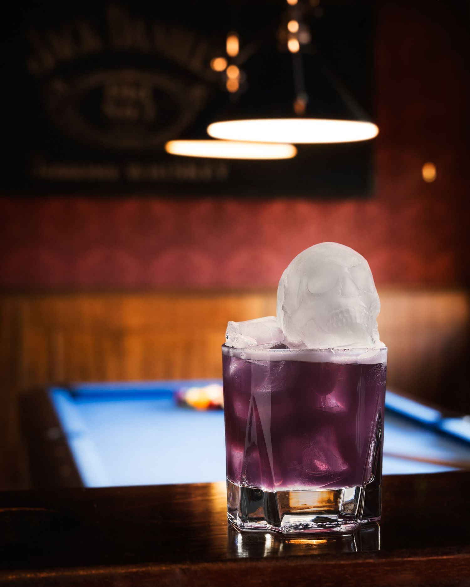 A purple cocktail with a large spherical ice ball served in a rocks glass on a wooden bar with ambient lighting.