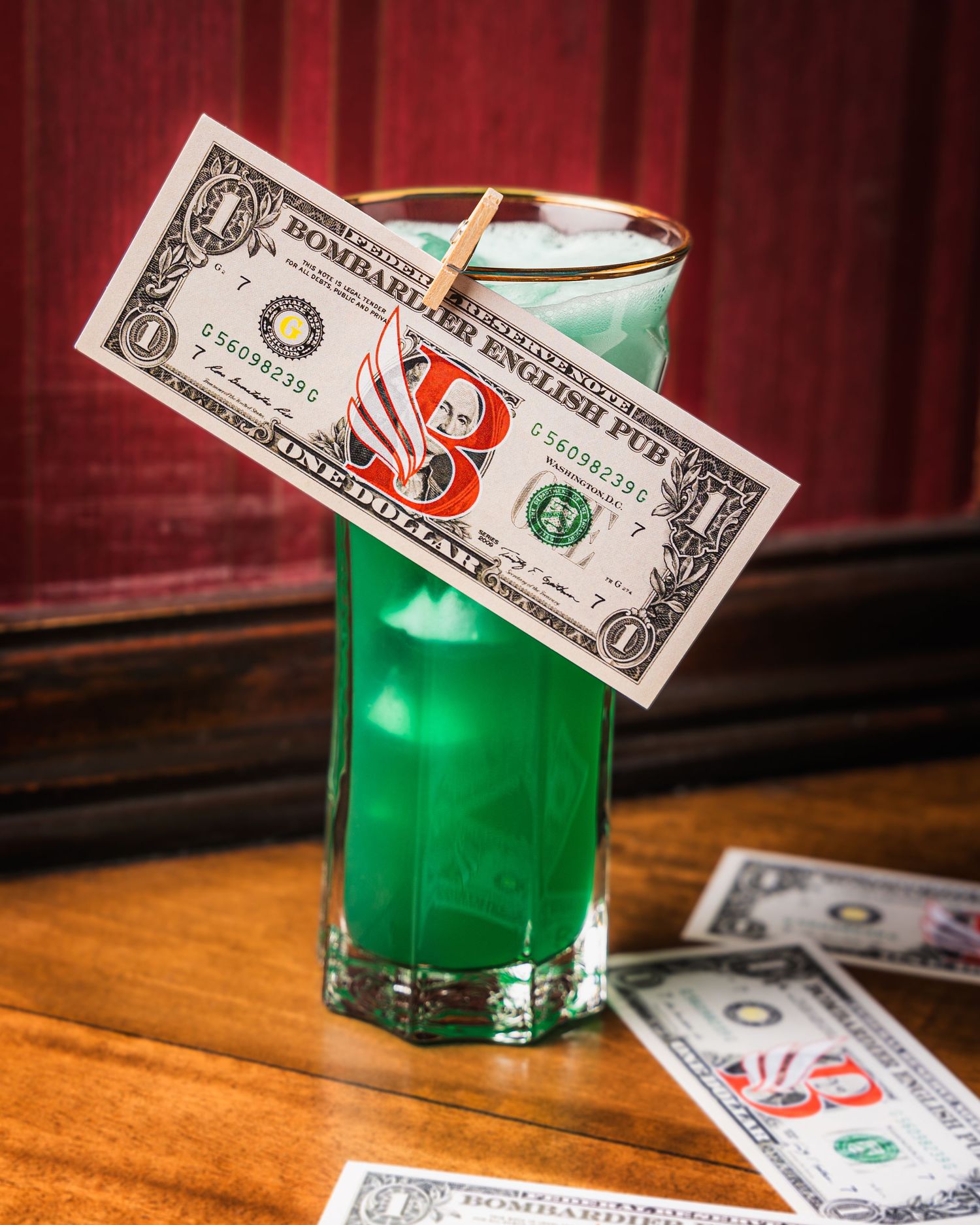 A green cocktail in a tall glass decorated with a one dollar bill on a wooden surface with more cash nearby.