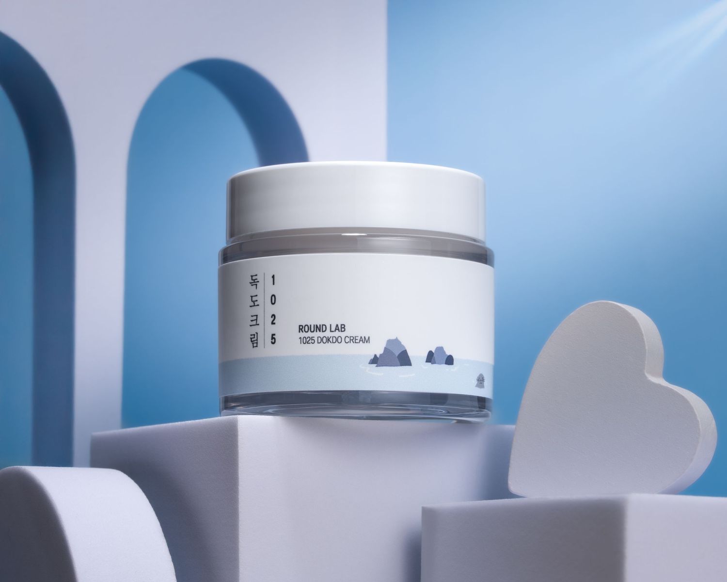 White skincare product jar displayed on geometric platforms with blue arches and white heart shapes in minimalist setting.