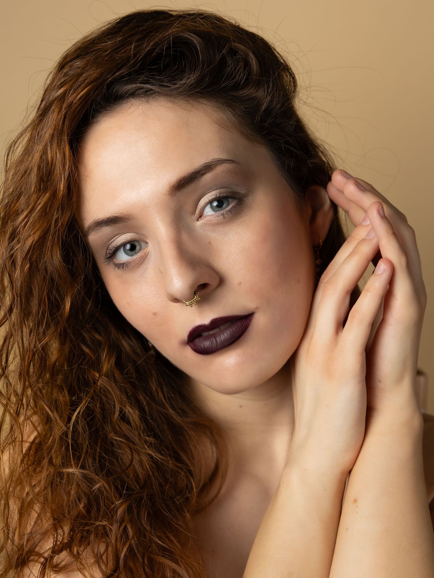 Dramatic beauty portrait featuring dark plum lipstick and curly auburn hair against a neutral beige background.