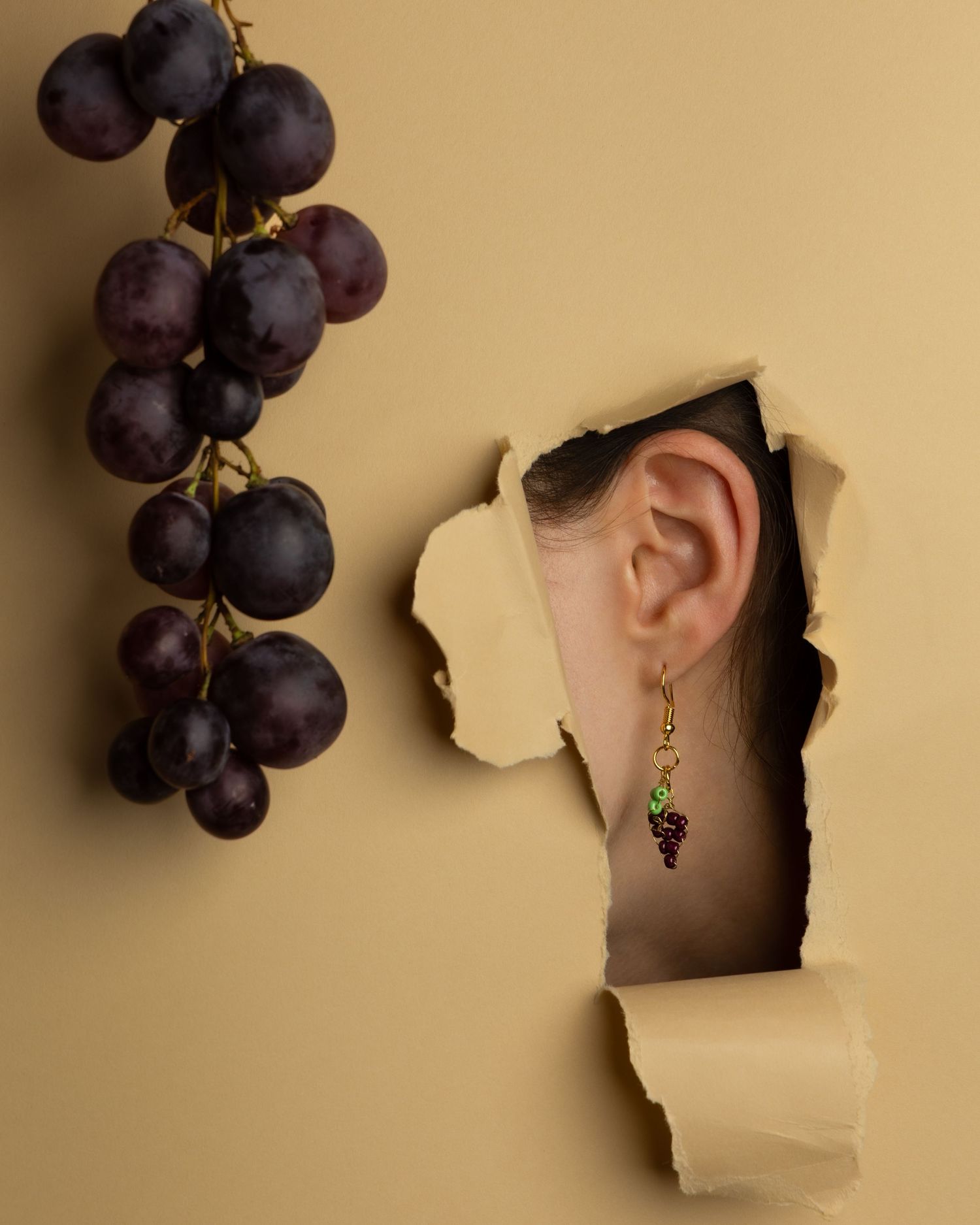 Artistic series showing purple grapes against beige background with shadows and dangling grape-shaped earrings.