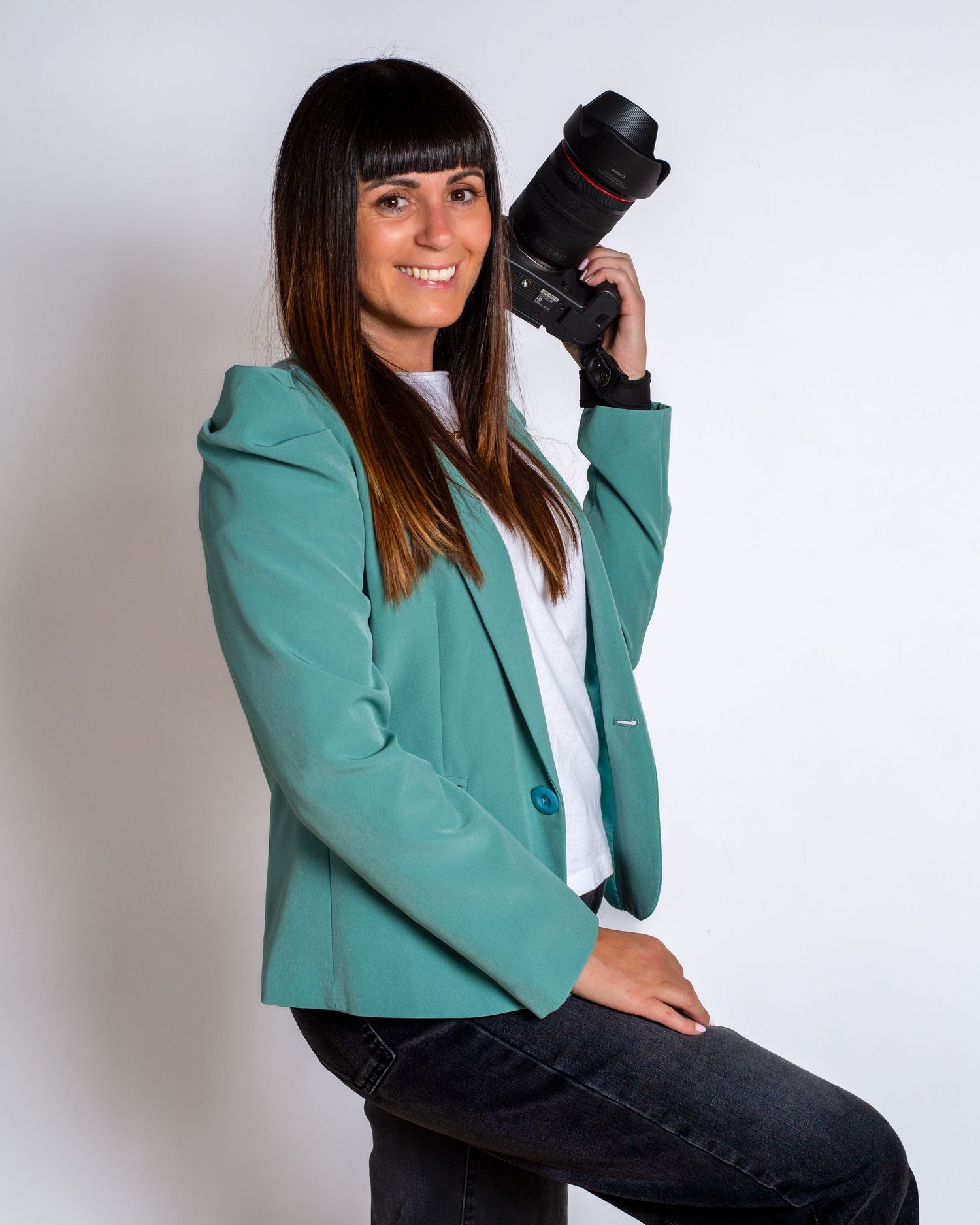 Professional photographer wearing a turquoise blazer holds a DSLR camera against a white studio background.