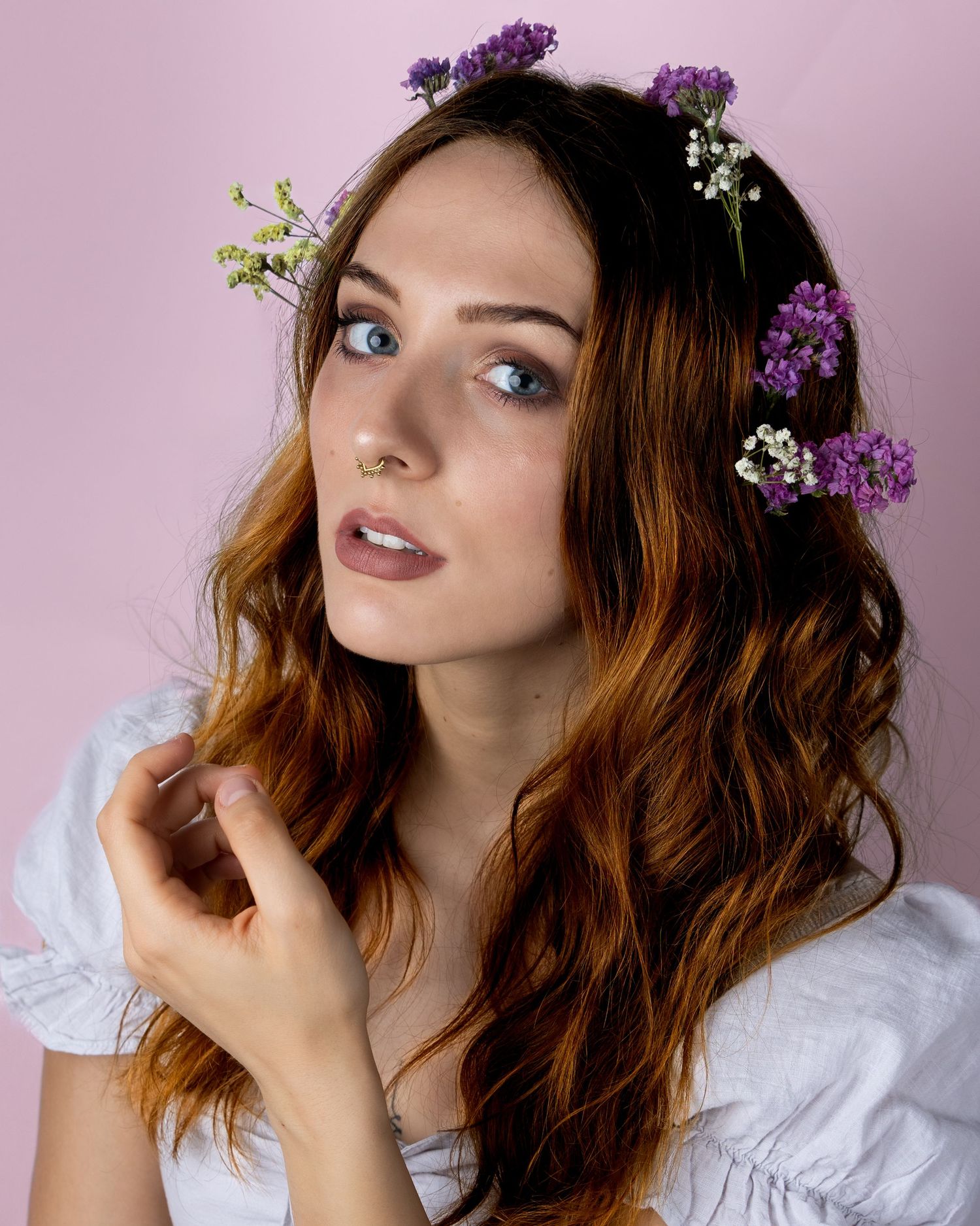 Ethereal portrait with flowing brown waves adorned with purple and white flowers against pink background in romantic style.