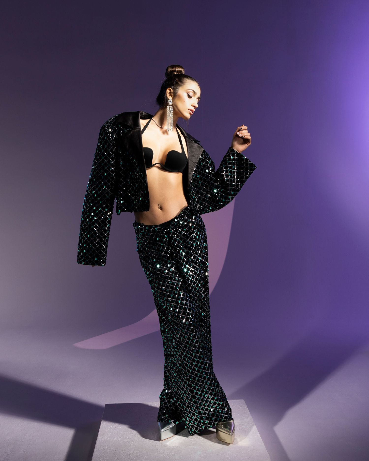 A dancer in a black sequined outfit with wide-leg pants and cropped jacket poses against a purple gradient background.