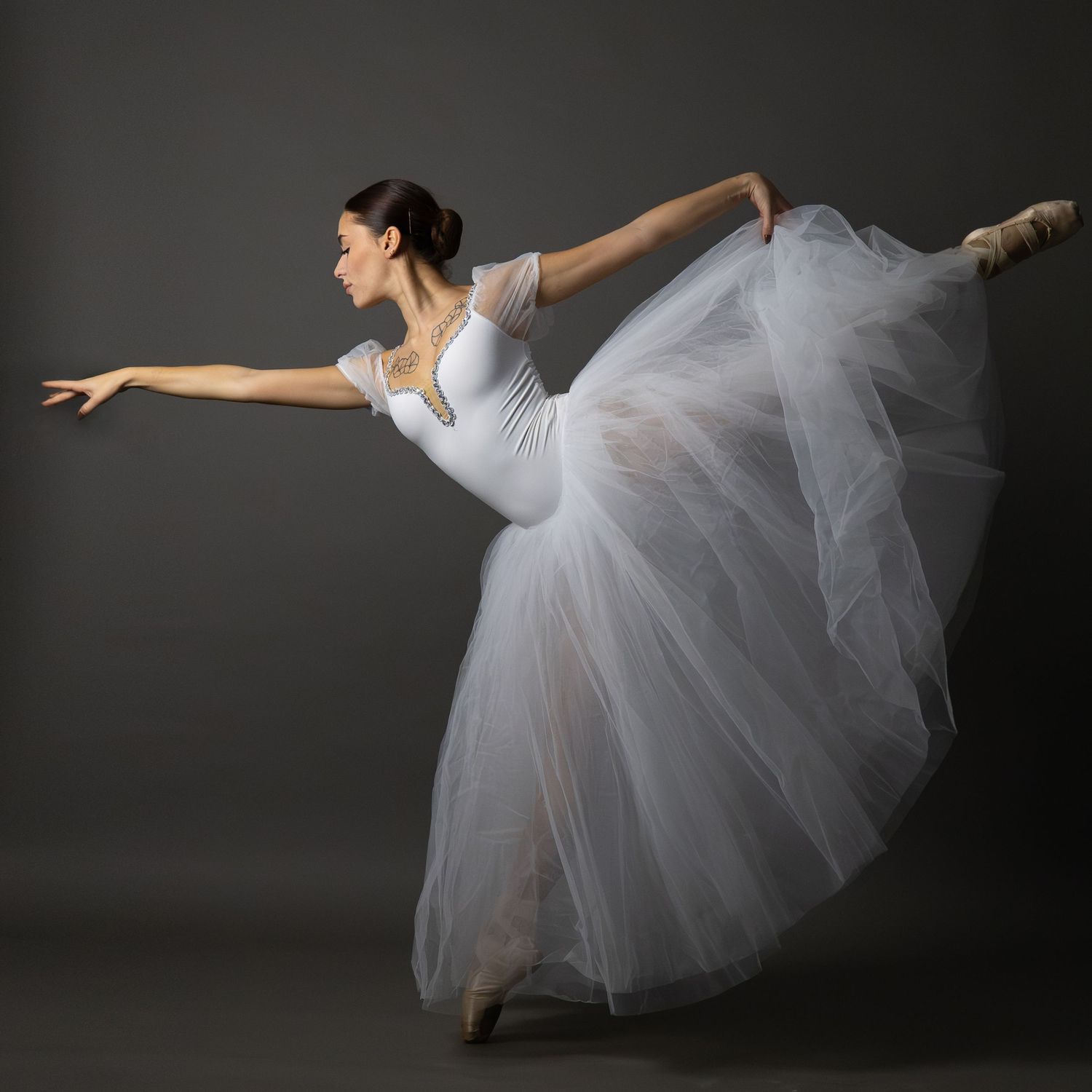 Ballet dancer in white tulle dress performs elegant pose against dark backdrop.