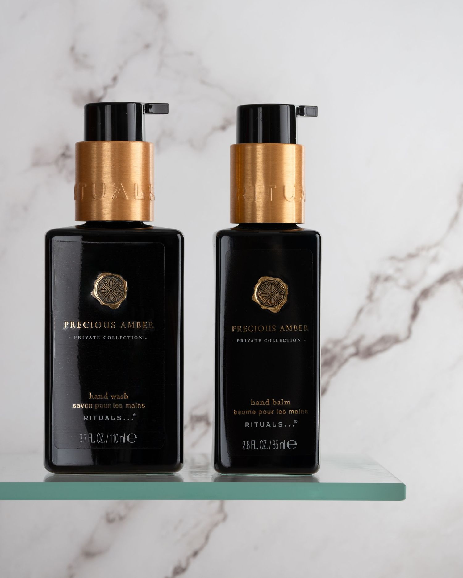 Two black luxury bath products with gold pump dispensers displayed on a glass shelf against white marble background.