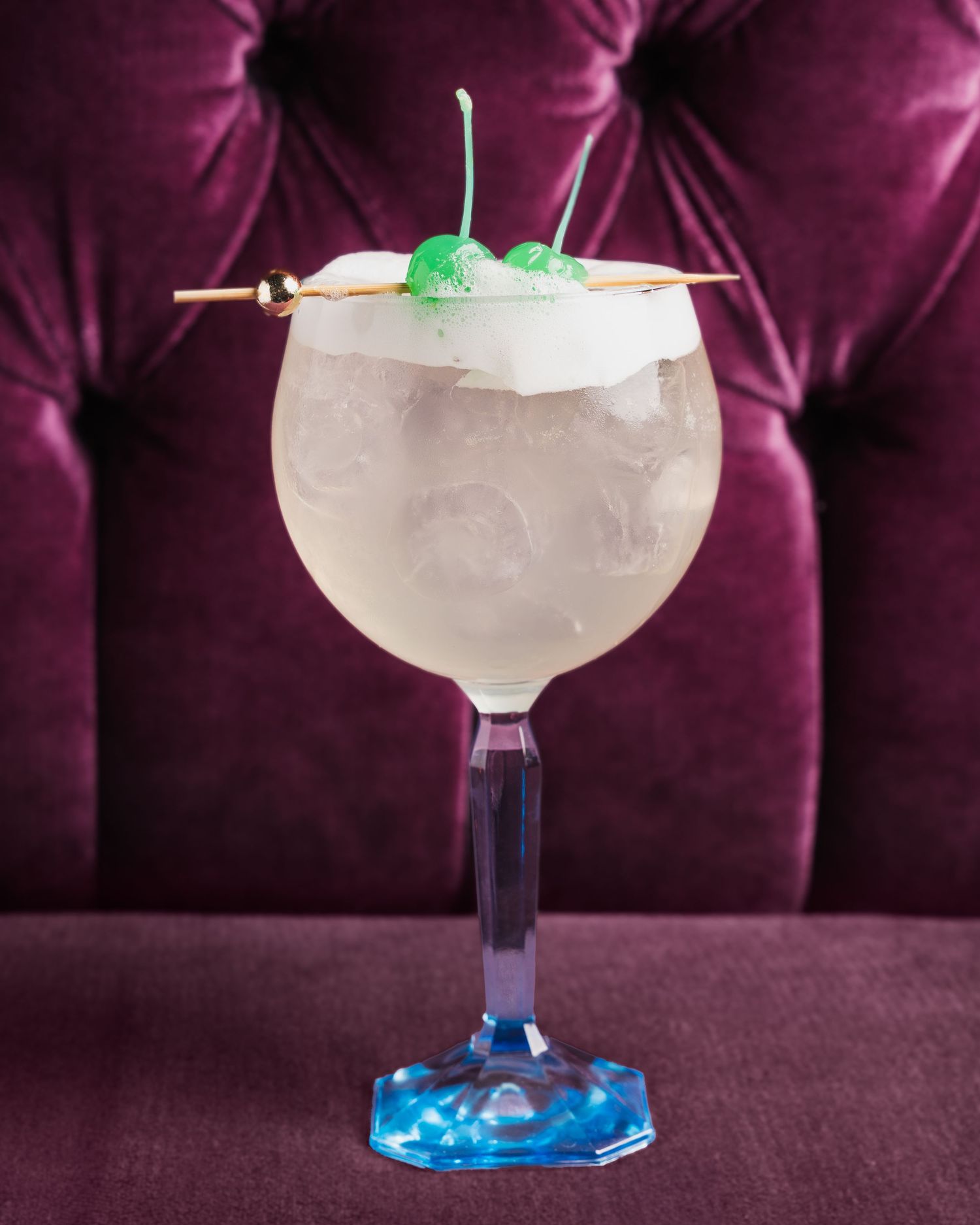 A frosty cocktail with green garnish served in a blue-stemmed goblet against a purple velvet backdrop.