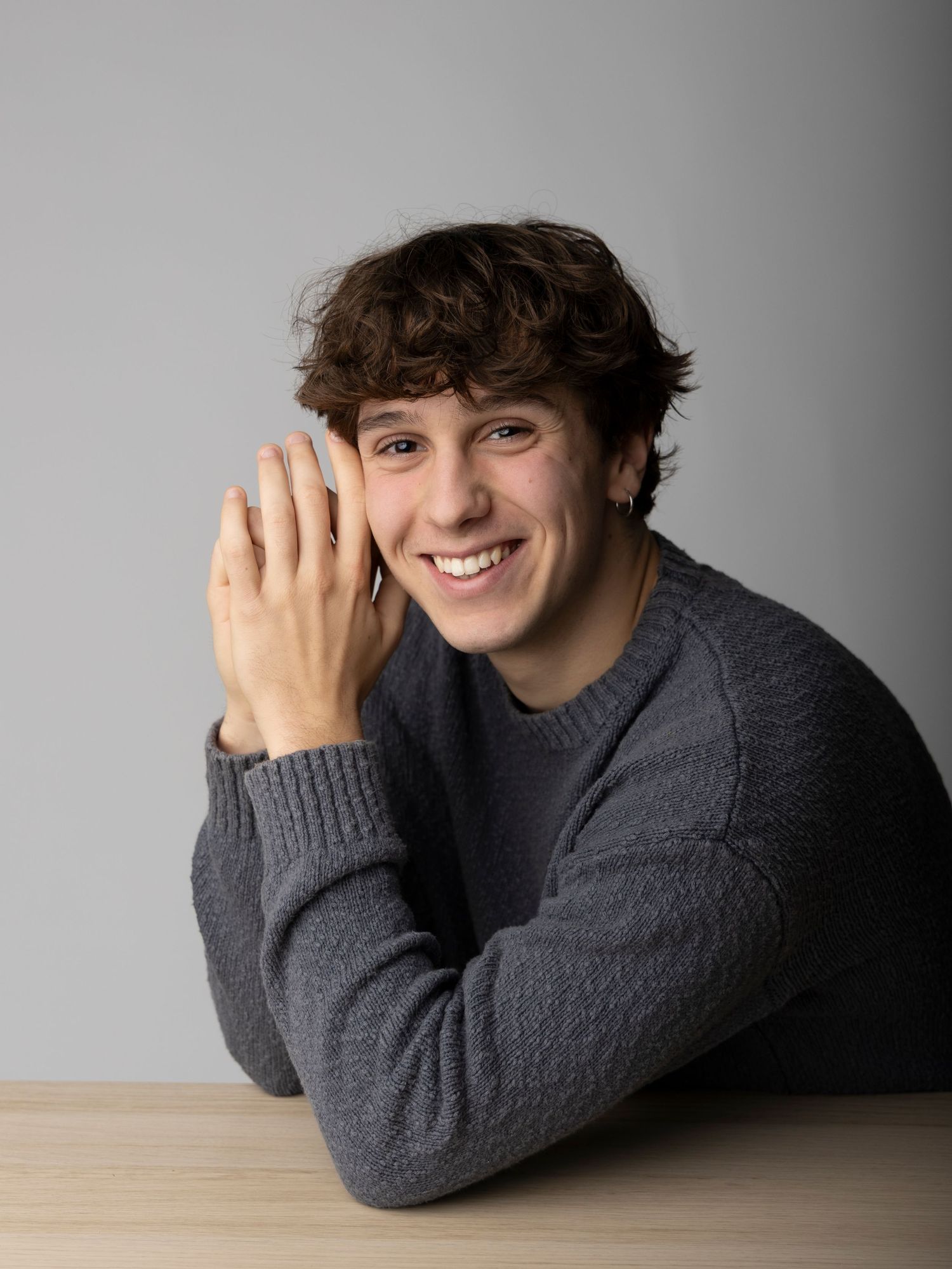 A person in a gray sweater lies on a wooden surface with a playful smile in a studio setting.