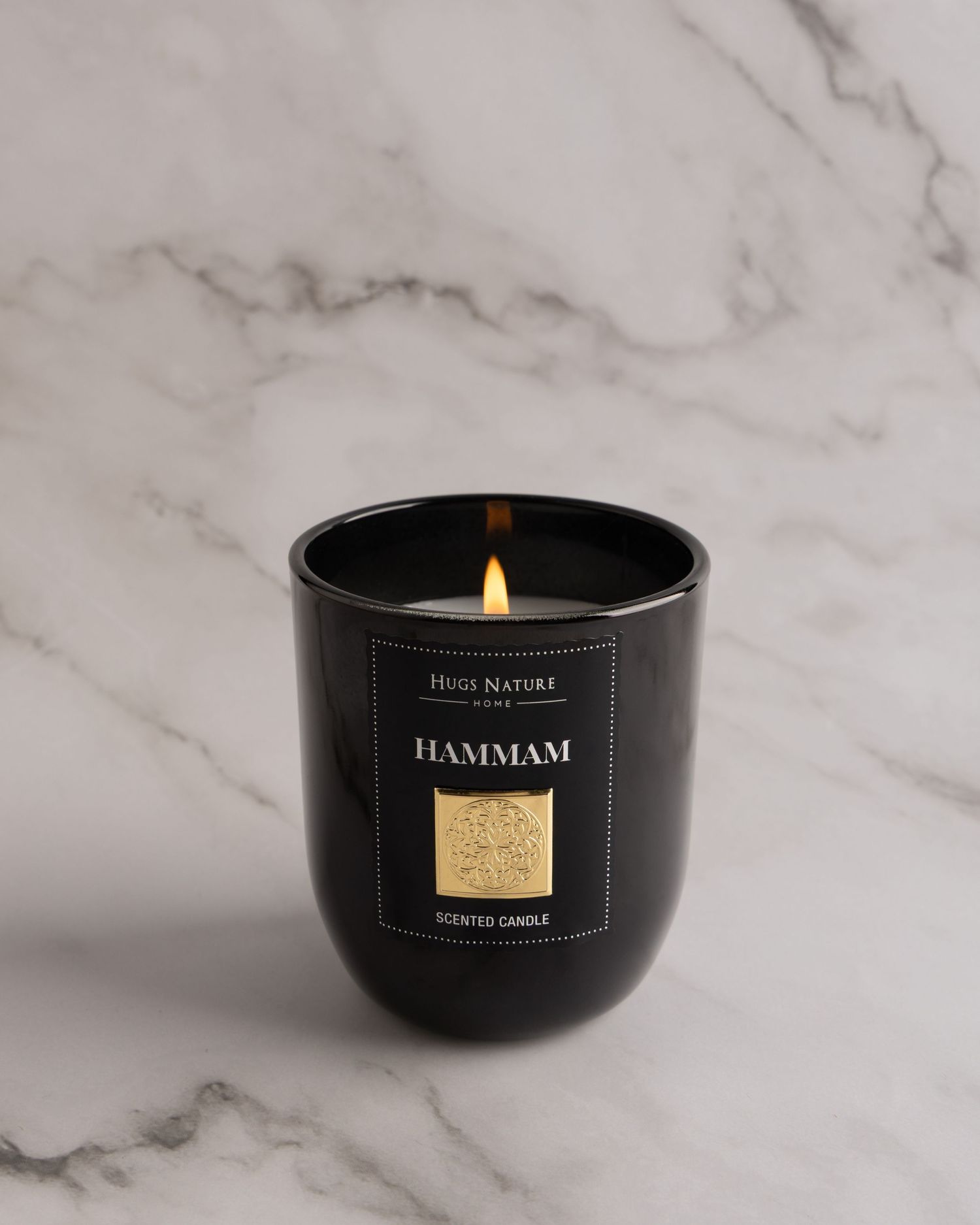Three black glass candle holders with gold labels sit on a white marble surface displaying luxury home fragrance products.