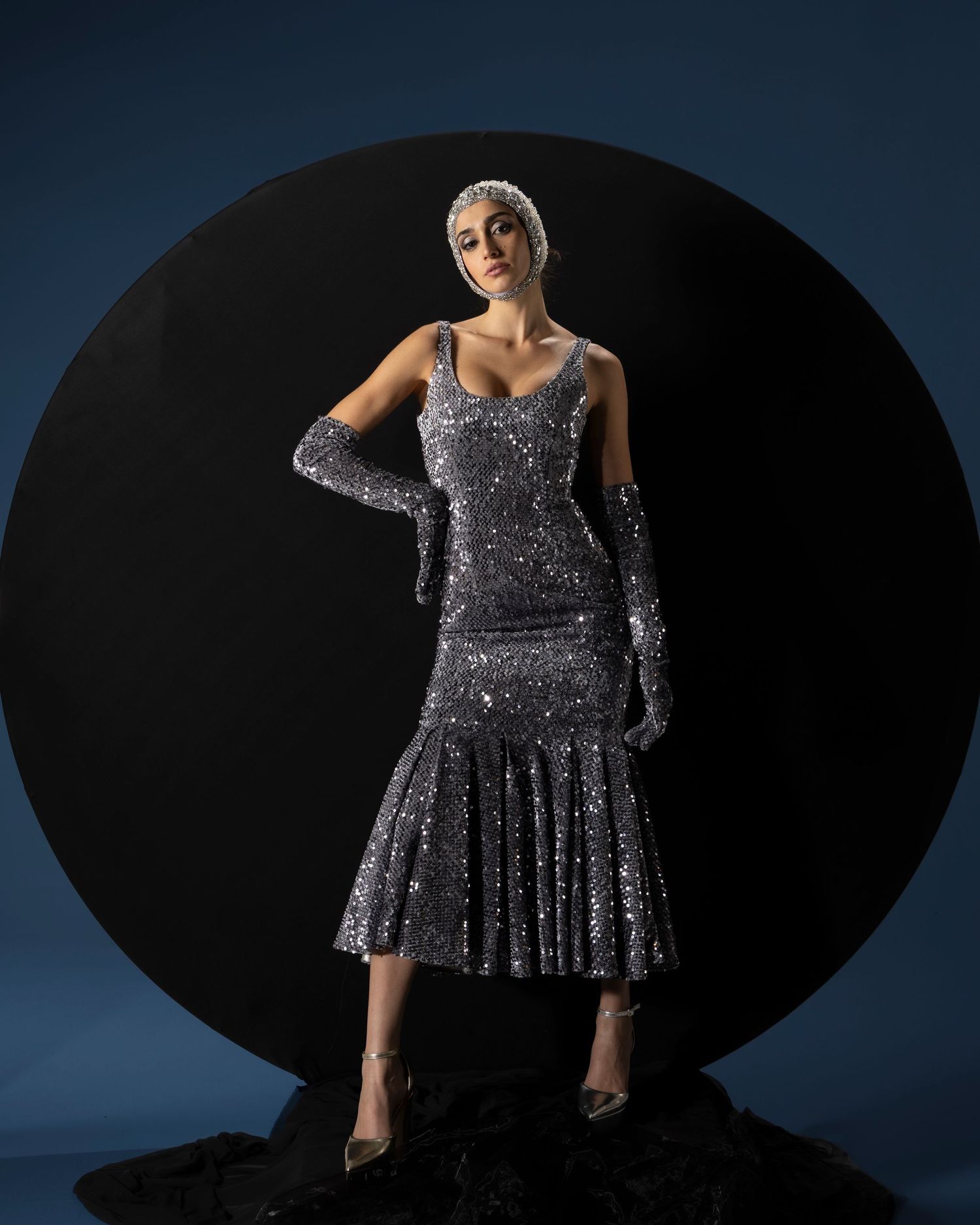 Figure in a glamorous silver sequined dress and long gloves poses dramatically against a dark circular backdrop.