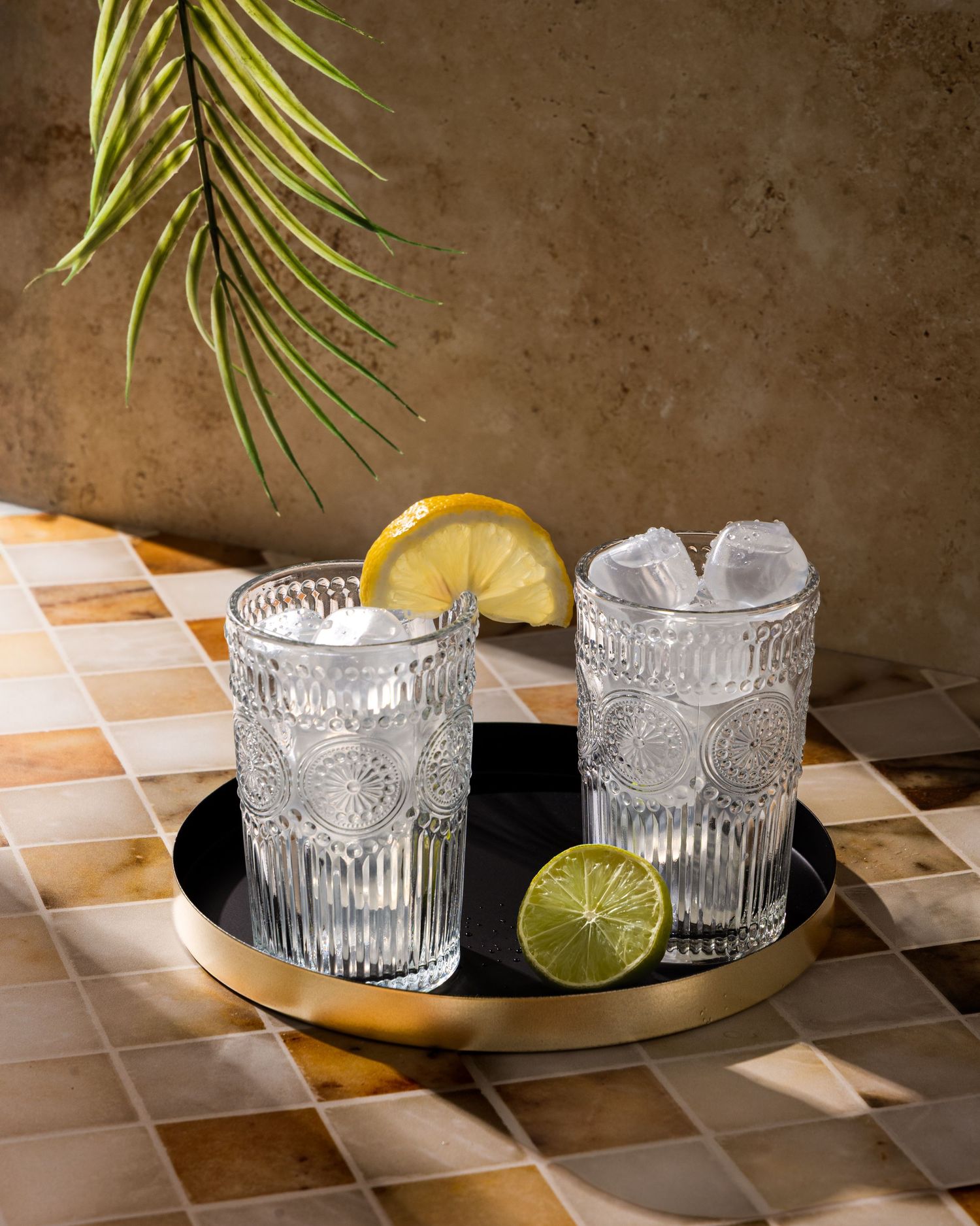 Two ornate glass cocktails with citrus garnishes on a gold serving tray against patterned tile and palm frond decor.