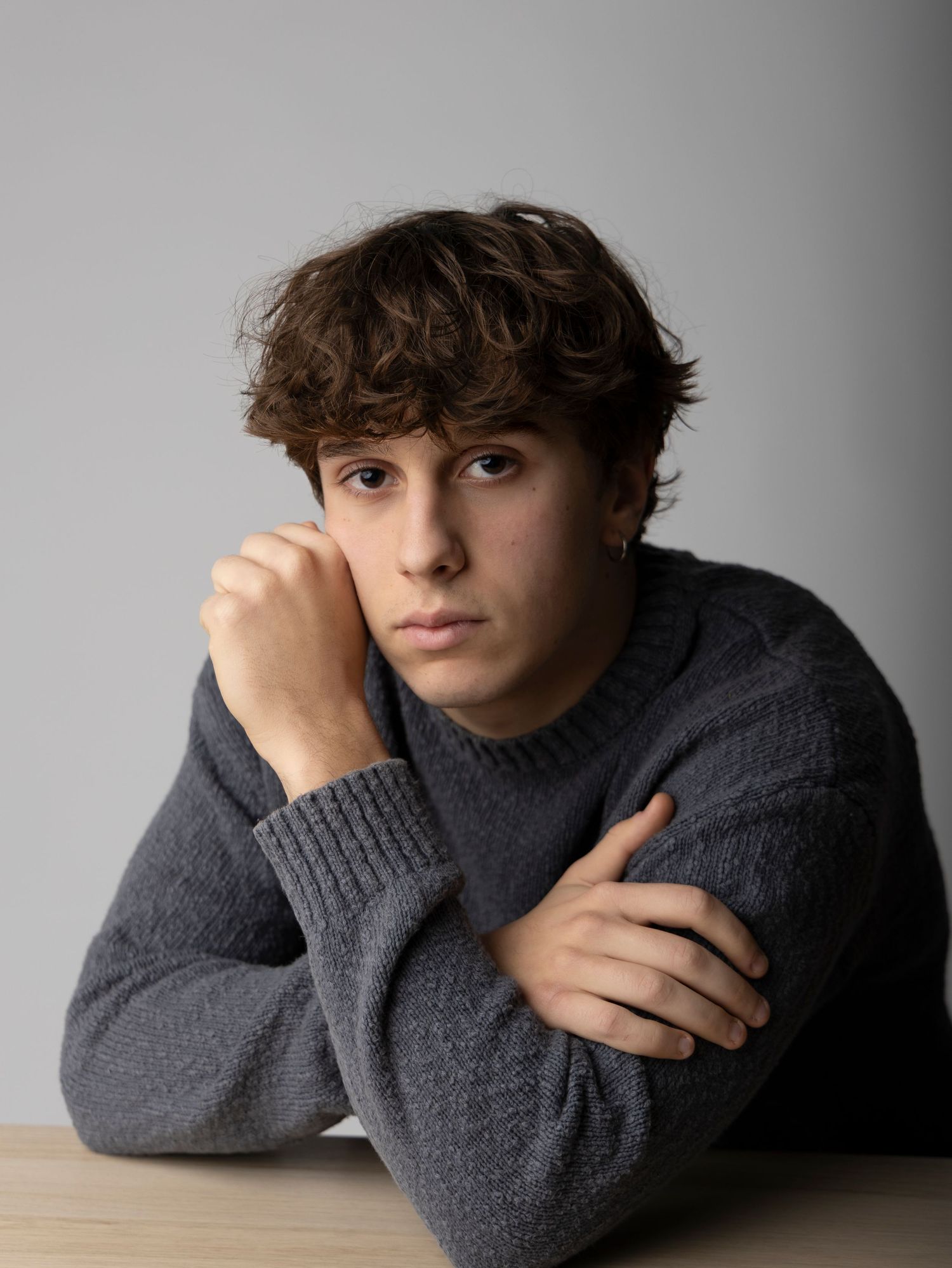 A figure in a dark gray sweater leans on a surface while posing against a light gray background in a series of photos.