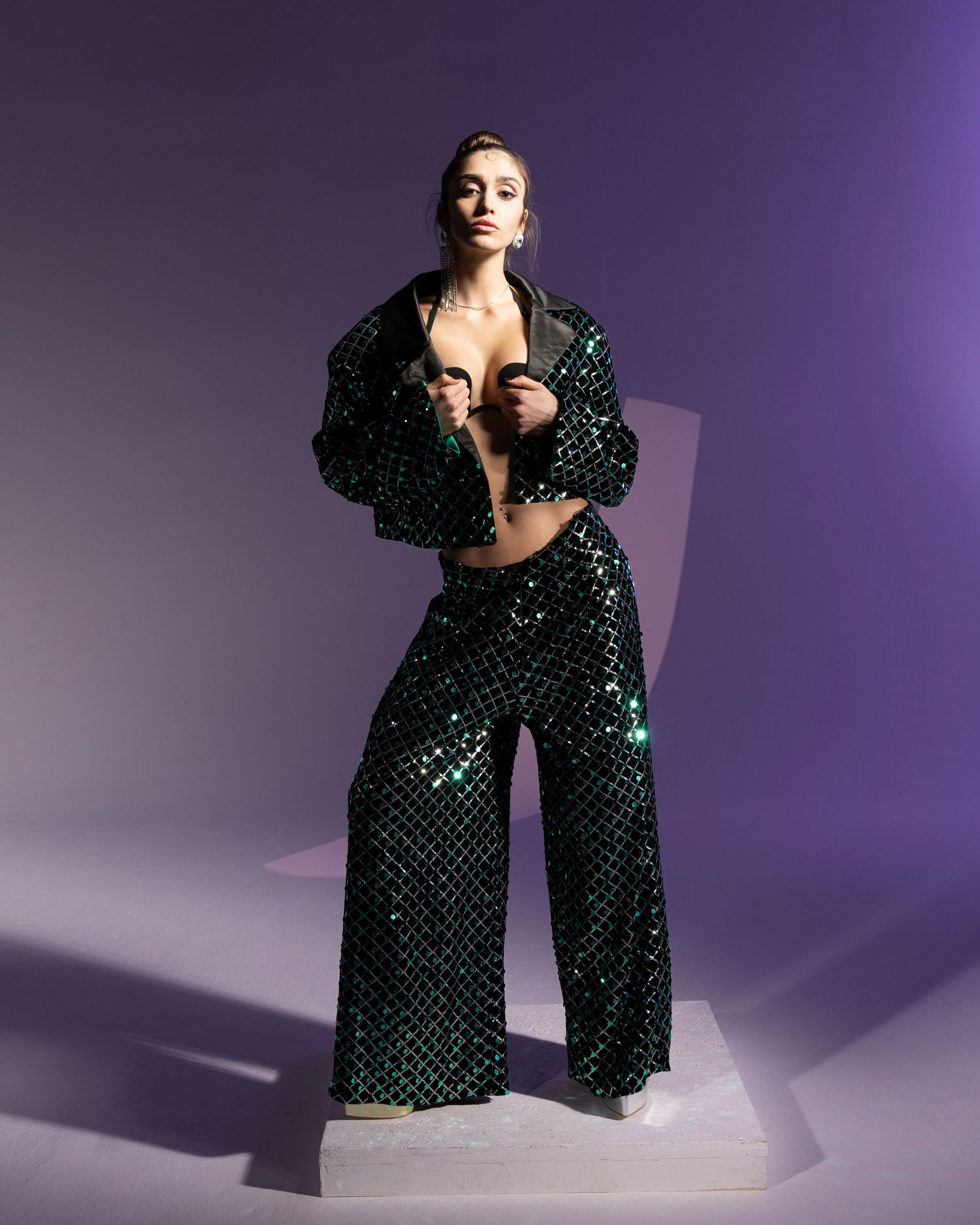 Fashion model poses in sparkling emerald green sequined pantsuit against purple backdrop with dramatic lighting.