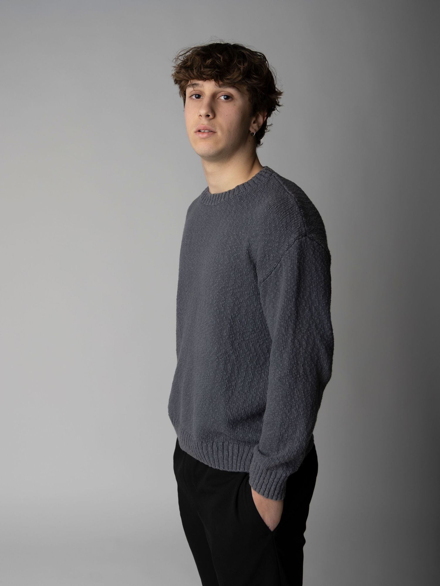 Studio portrait of someone in a textured gray sweater and dark pants against a neutral background.