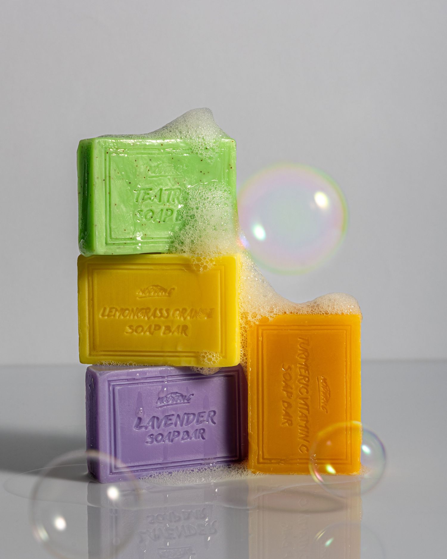 Colorful artisanal soap bars in purple, green, and orange arranged with iridescent bubbles on a light gray background.