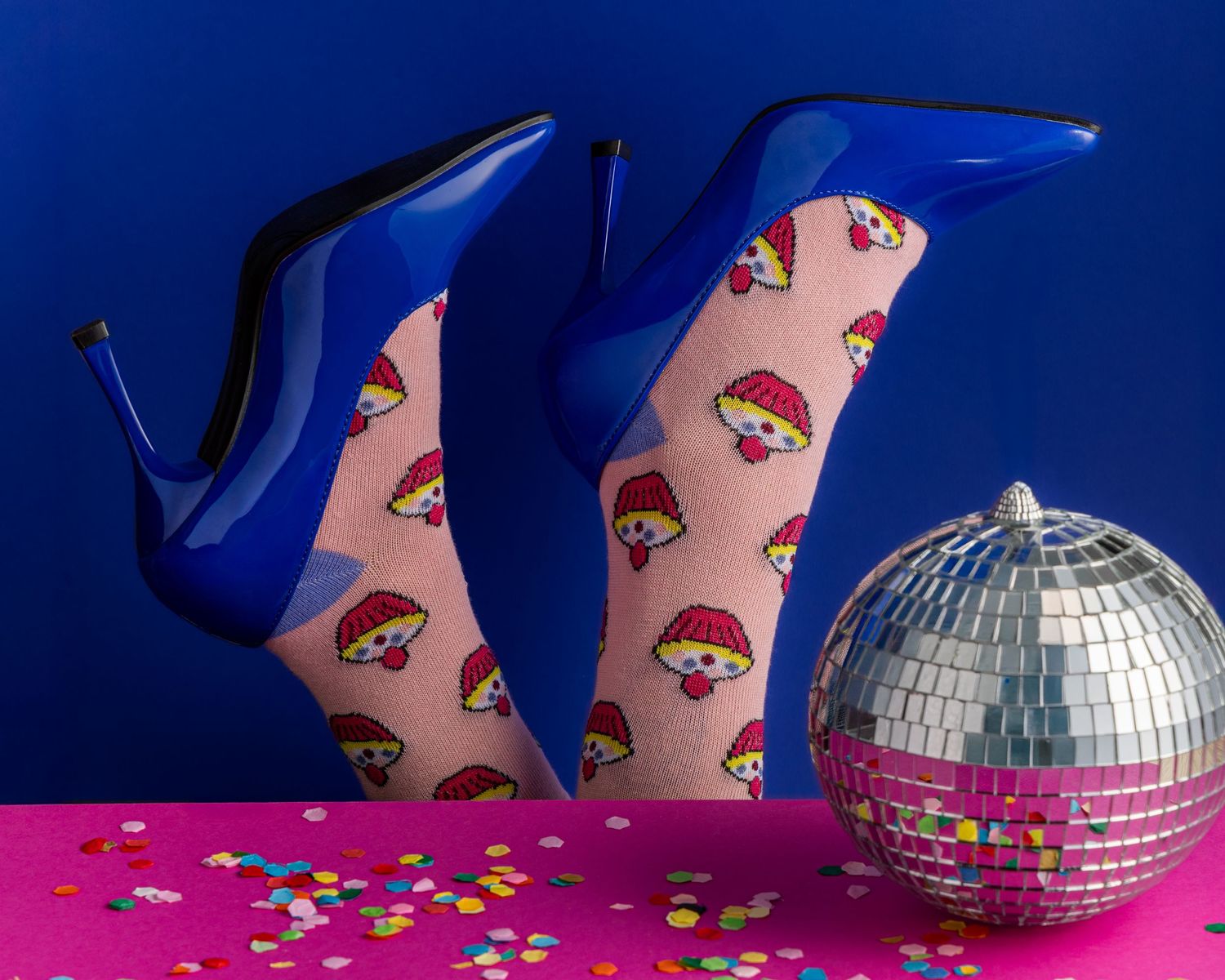 Blue high heel shoes with red lip print pattern next to a disco ball on pink surface with scattered confetti.