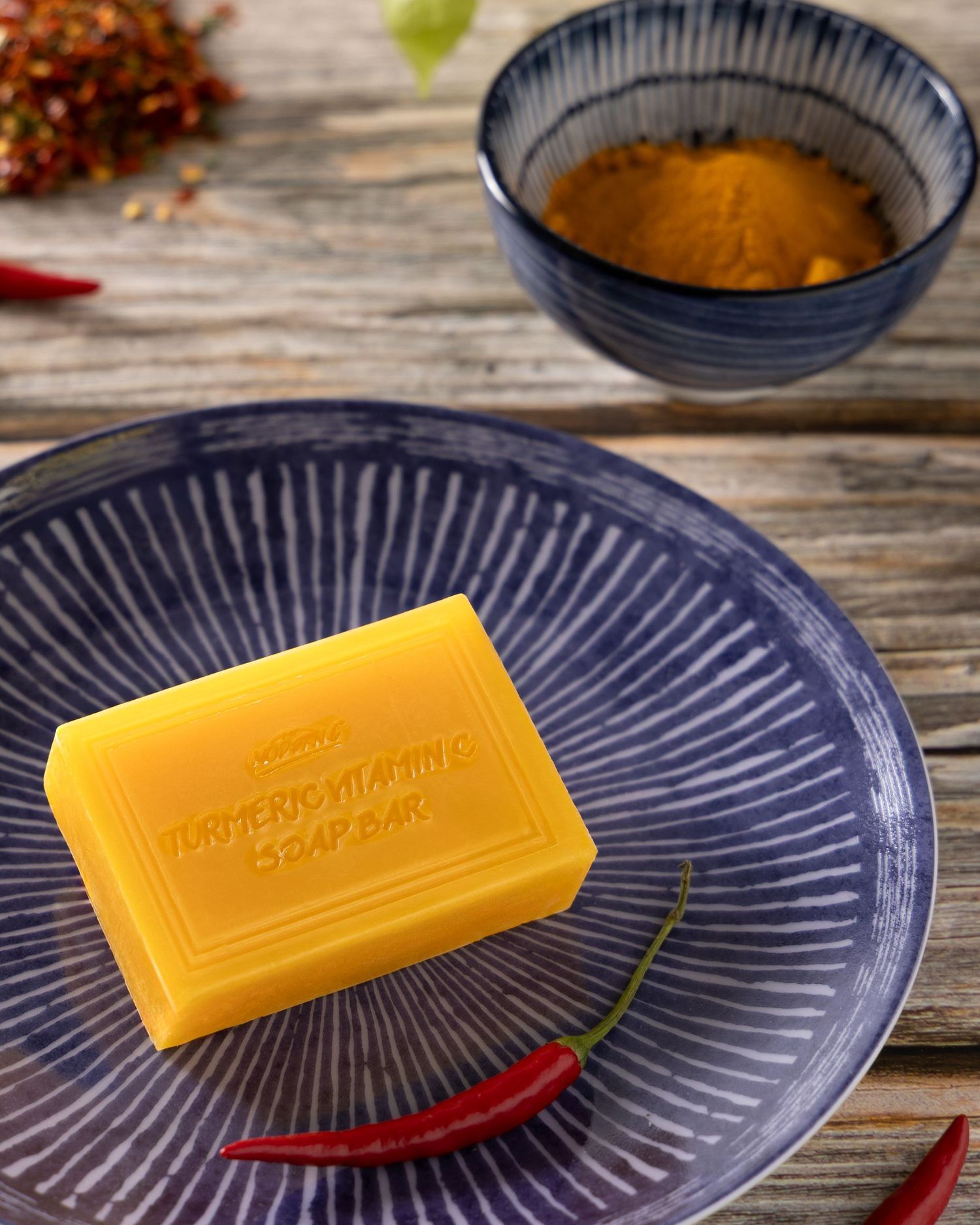 Yellow turmeric soap bar on blue plate with red chili peppers and turmeric powder in bowl on wooden surface.