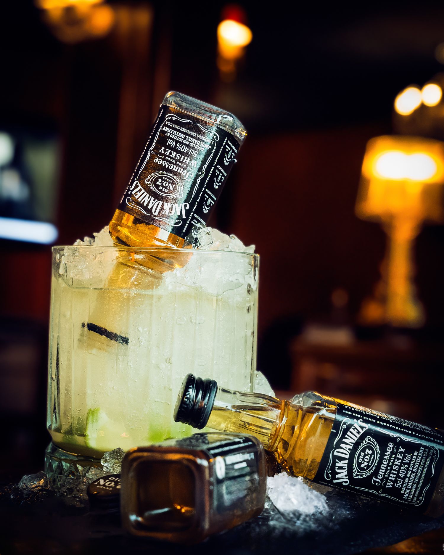 Two Jack Daniel's whiskey bottles perched on a cocktail glass with ice and lime in a dimly lit bar setting.