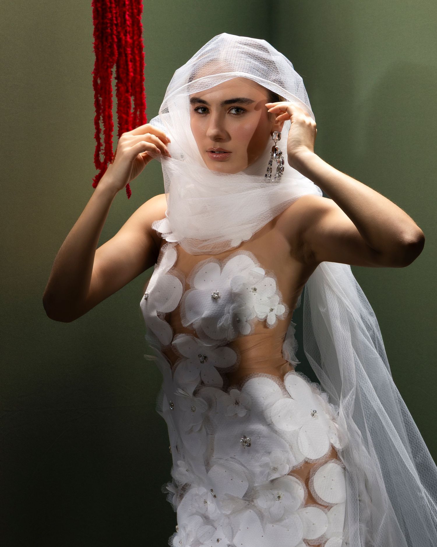 A figure in a white avant-garde bridal gown with sheer veil and floral appliques poses against a dark green background.