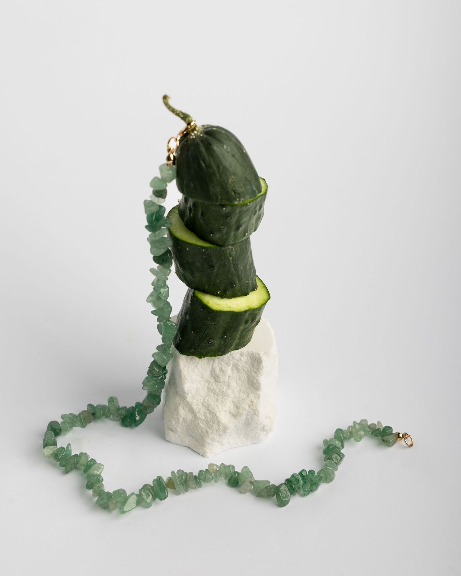 Beaded green gemstone necklace with rough cut stones drapes across white marble display blocks with green vegetables.