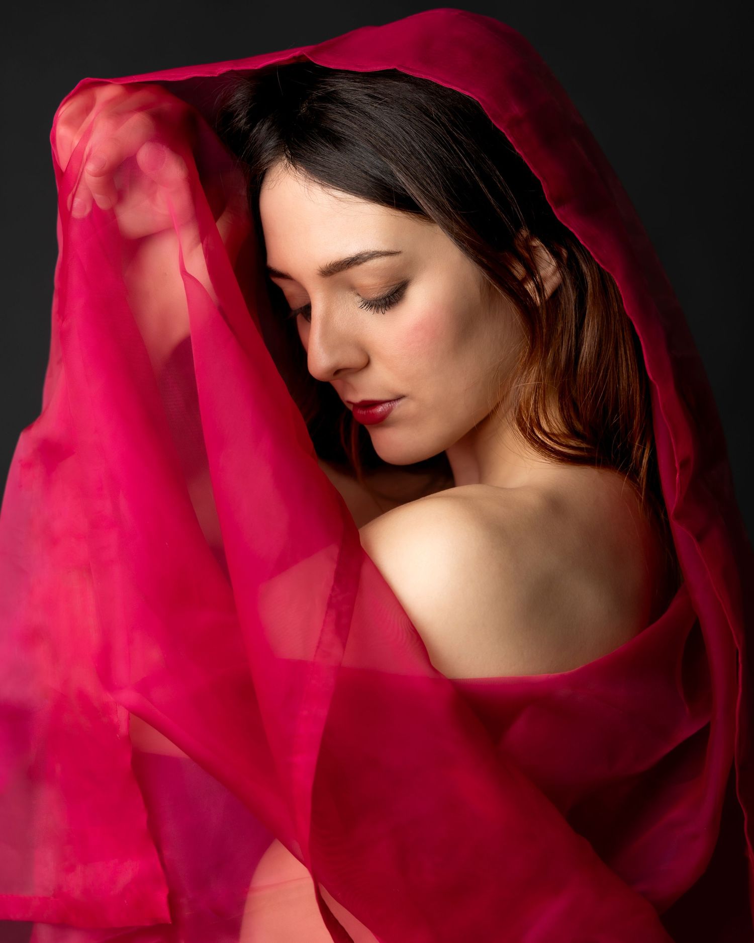 A woman draped in vibrant red silk fabric poses gracefully with eyes closed against a dark background.