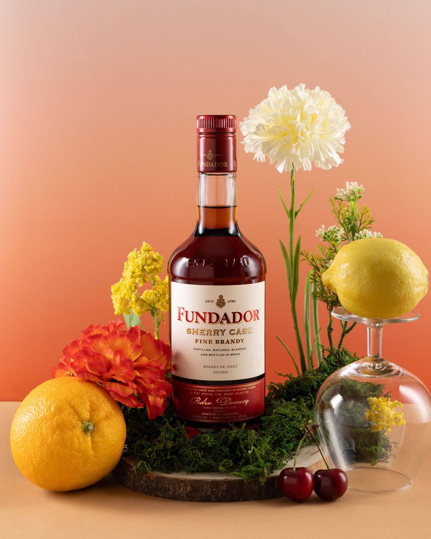 A bottle of Fundador brandy displayed with oranges, flowers, and cherries against a peach-colored background.