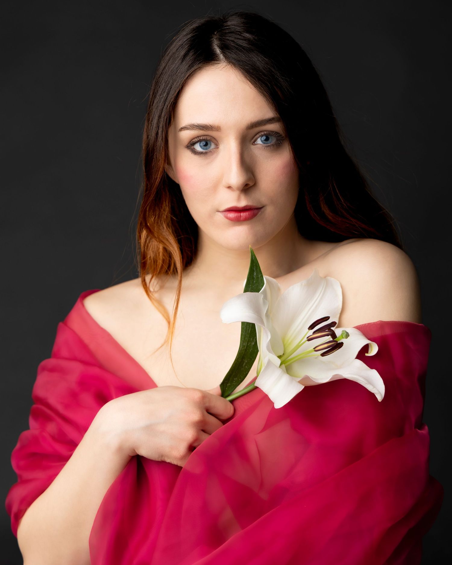 Young woman with blue eyes holding a white lily, draped in a vibrant pink fabric against a dark background.