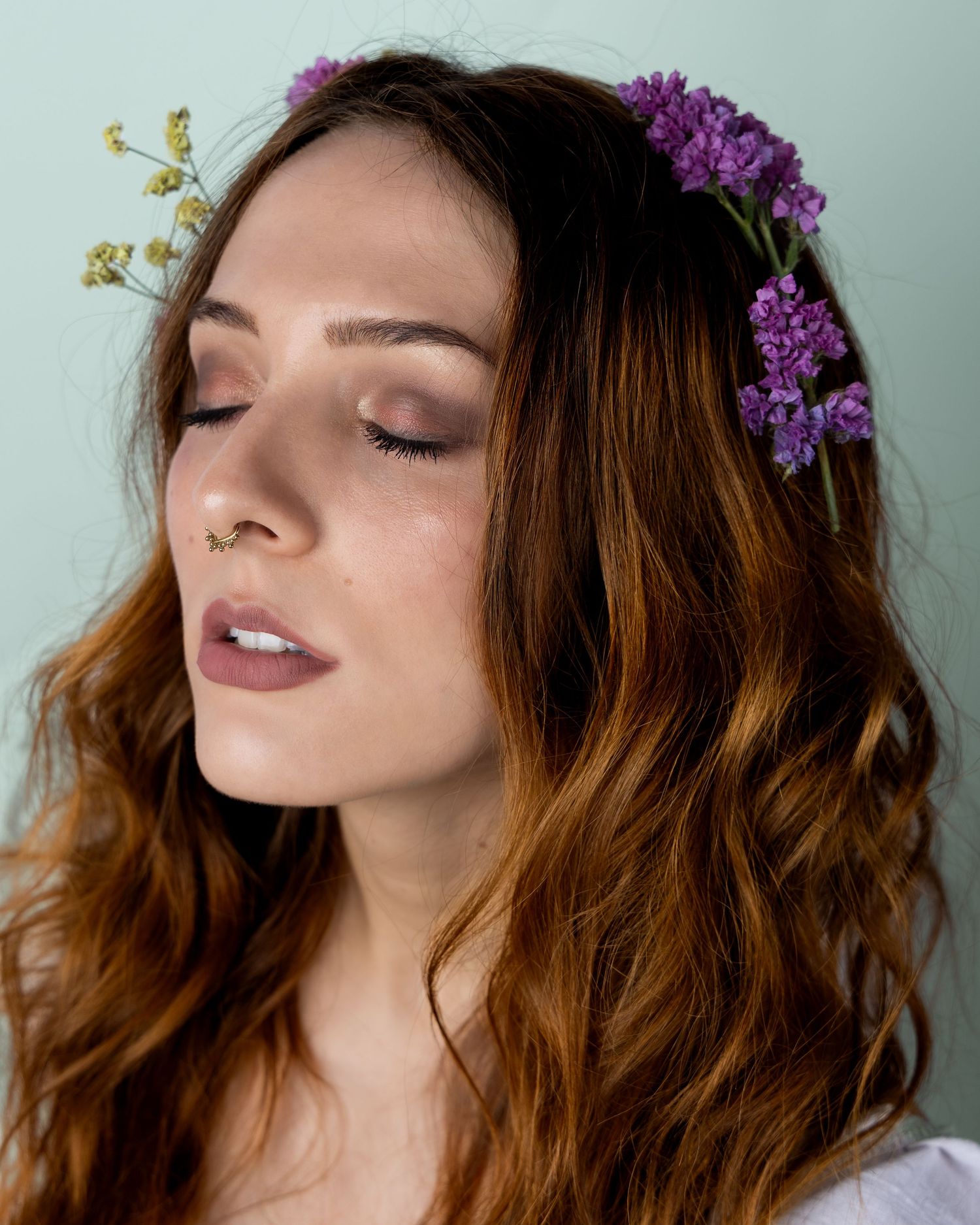 Dreamy ethereal portrait with purple flowers adorning wavy hair and natural makeup in soft light.